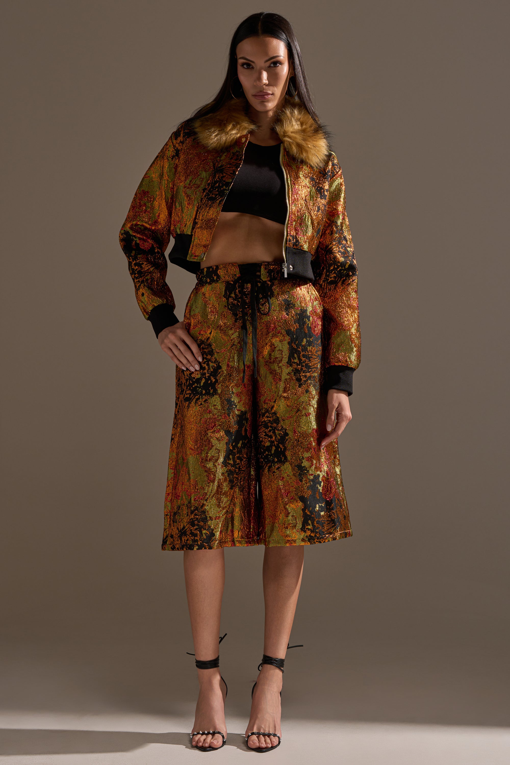 A woman stands confidently in the TWIN FLAME BROCADE BOMBER with a faux fur collar, paired with a black crop top, warm-toned patterned high-waisted skirt, and black strappy heels.