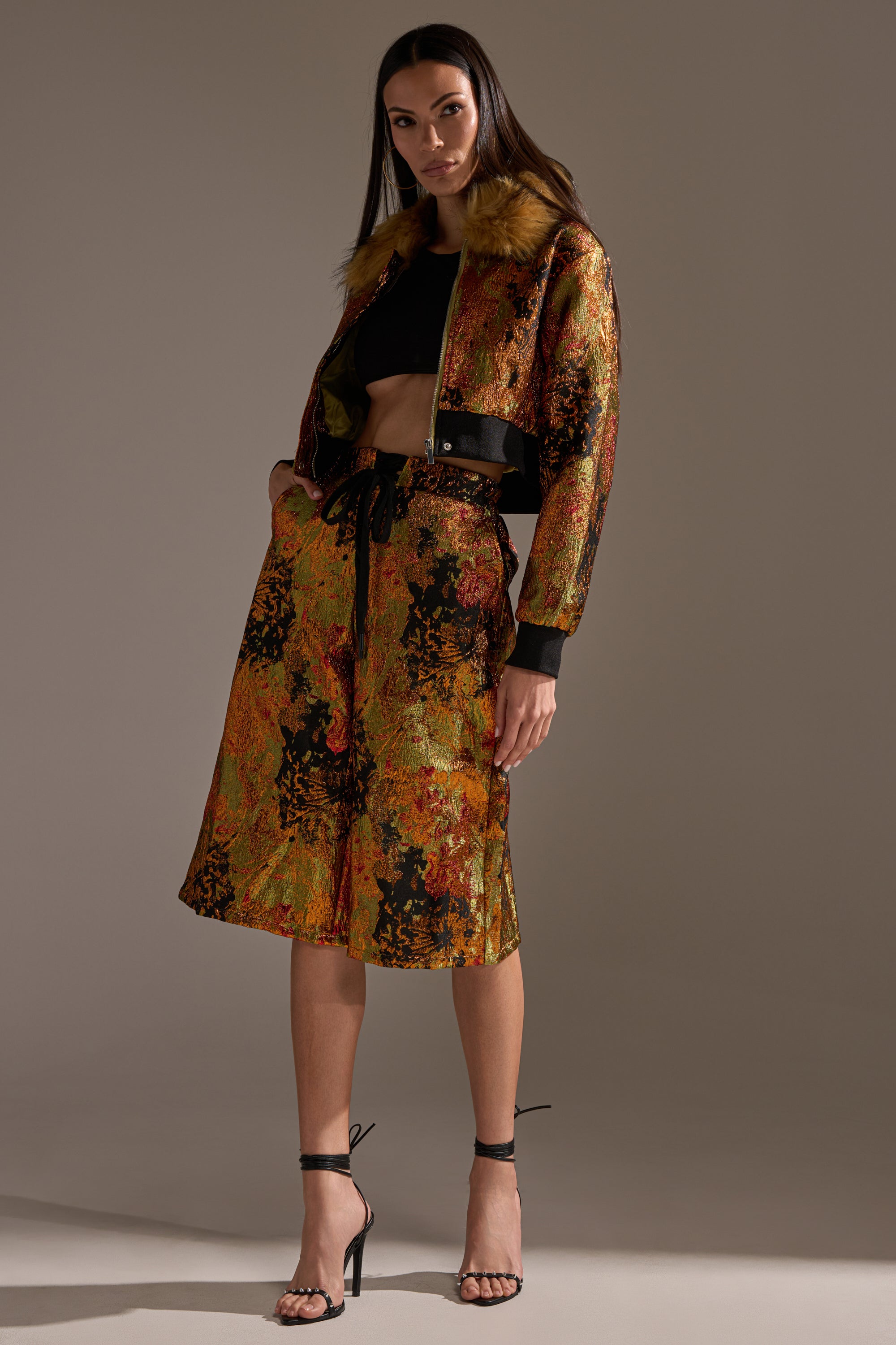 A woman stands confidently in the TWIN FLAME BROCADE BOMBER with faux fur collar, paired with a matching knee-length skirt, black crop top, and strappy black heels against a plain background.