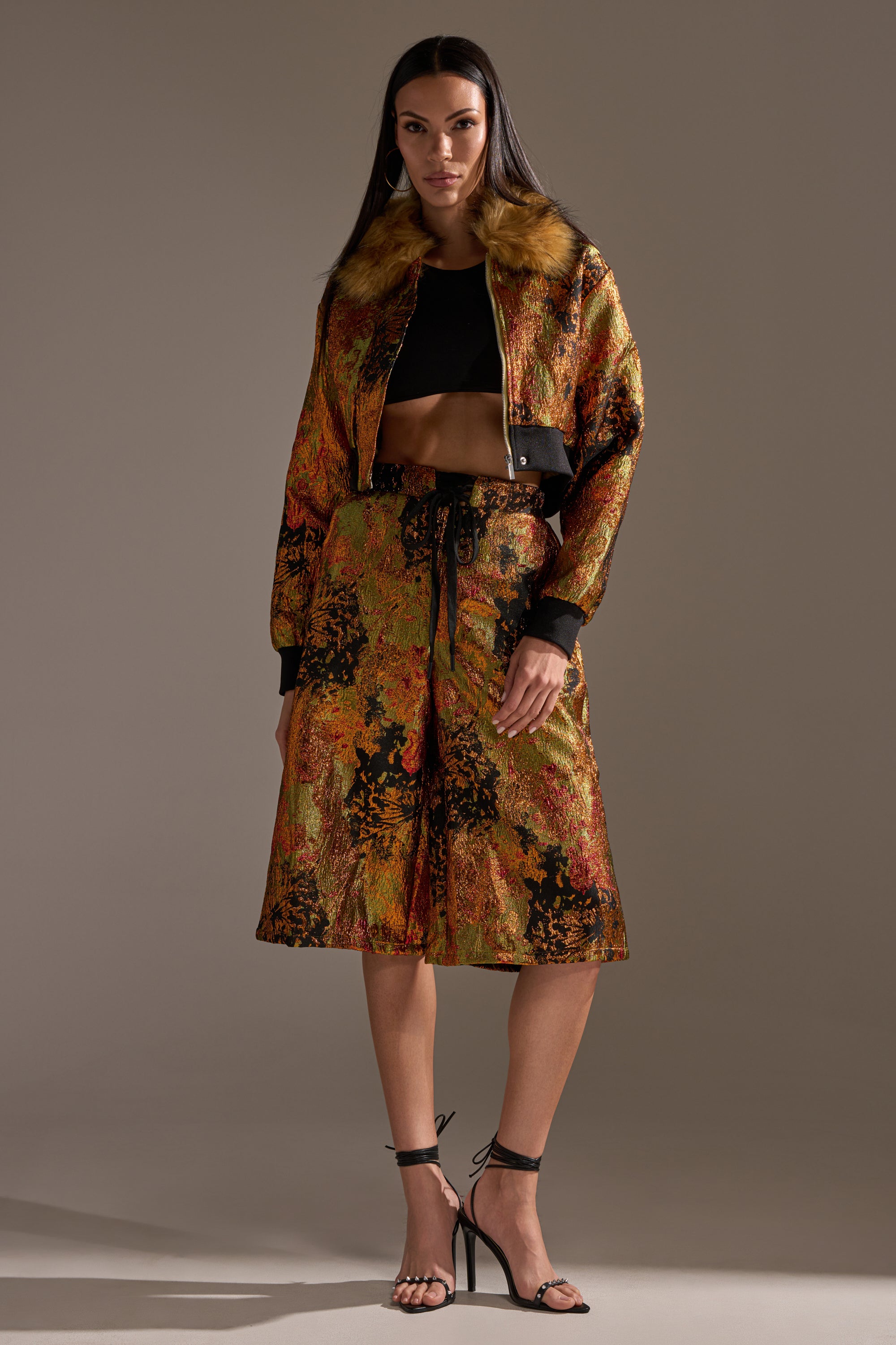 A woman wears the TWIN FLAME BROCADE BOMBER with a faux fur collar, styled with a black crop top, matching knee-length skirt, and black strappy heels against a simple neutral background.