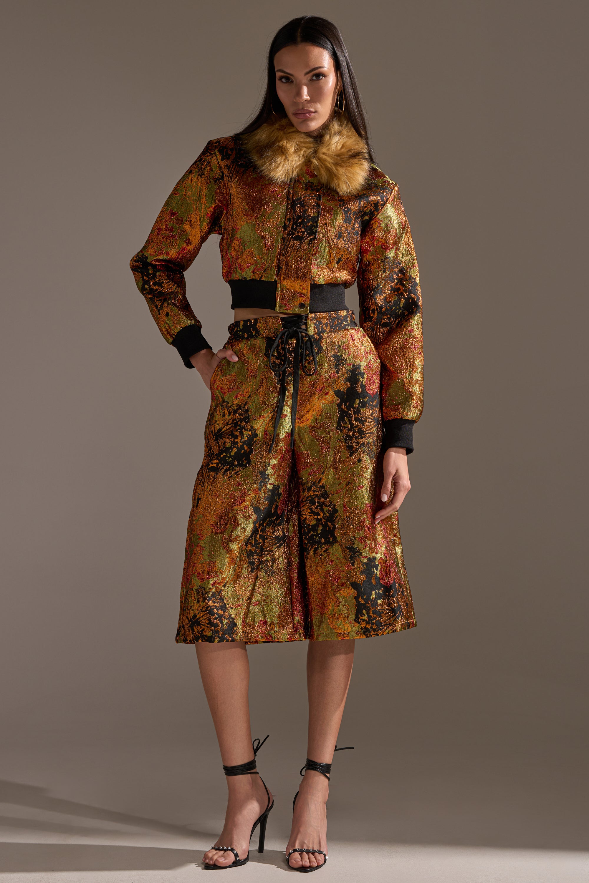 A woman with long dark hair models the TWIN FLAME BROCADE BOMBER in a studio, featuring a metallic finish, cropped fit, and faux fur collar. She pairs it with a matching knee-length skirt and black high-heeled sandals.