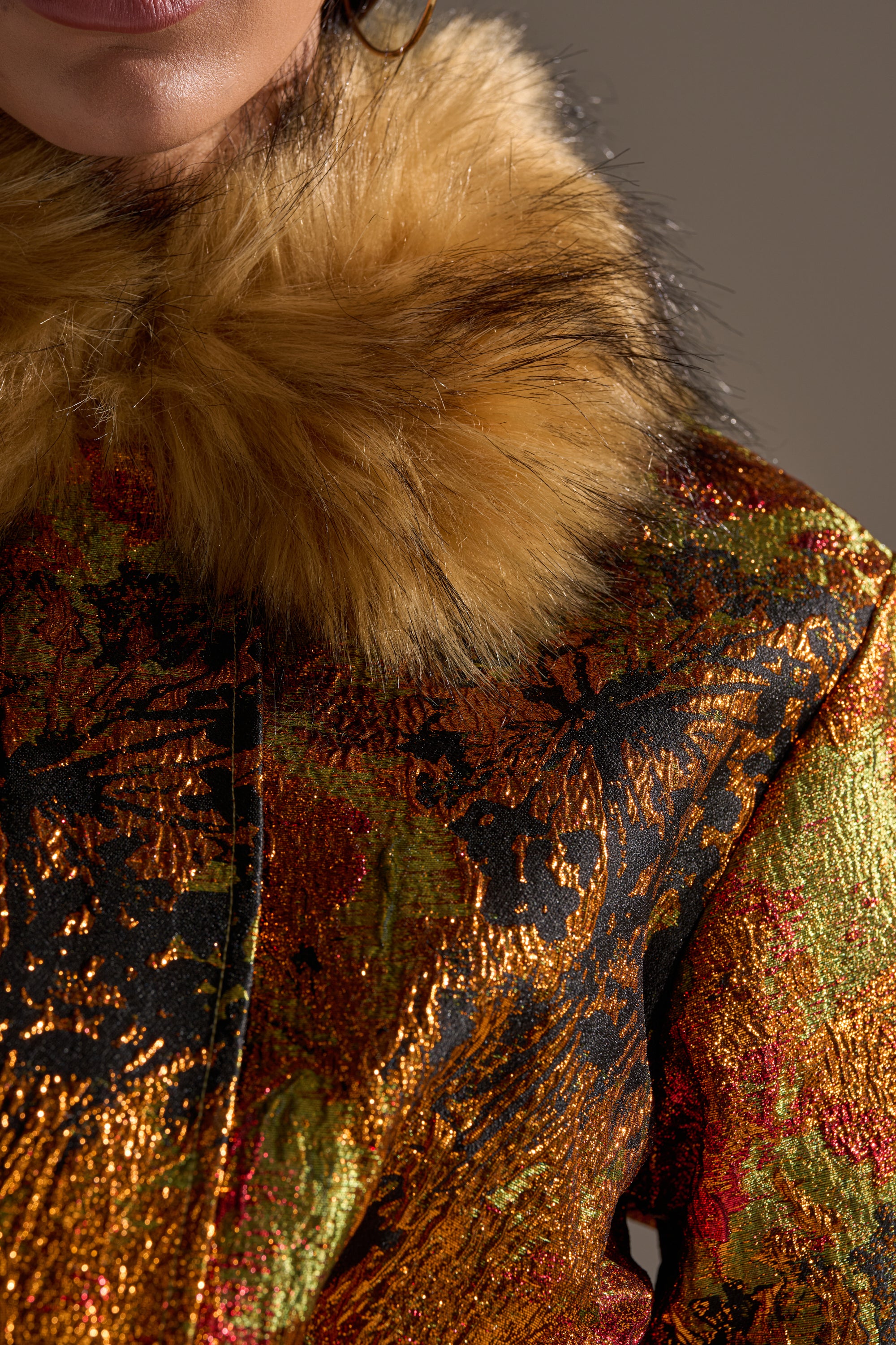 A close-up of a person wearing the TWIN FLAME BROCADE BOMBER, featuring a cropped fit, metallic gold and black pattern, and a plush faux fur collar in golden brown and black. Only the lower face and neck are partially visible.