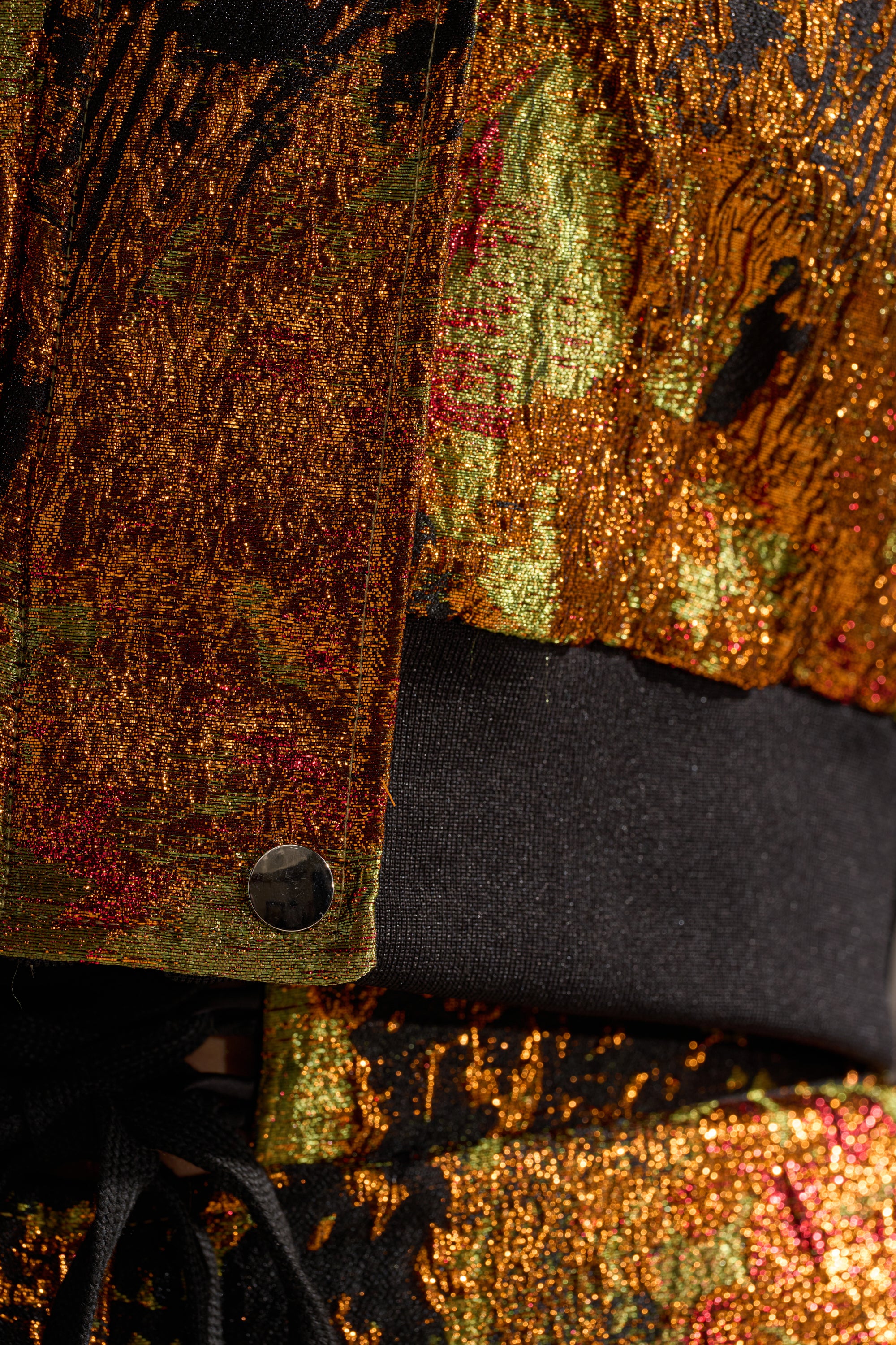 Close-up of the TWIN FLAME BROCADE BOMBER, featuring textured gold, orange, and green threads, a black waistband, round metallic button, and shiny brocade fabric in a cropped fit for modern style.
