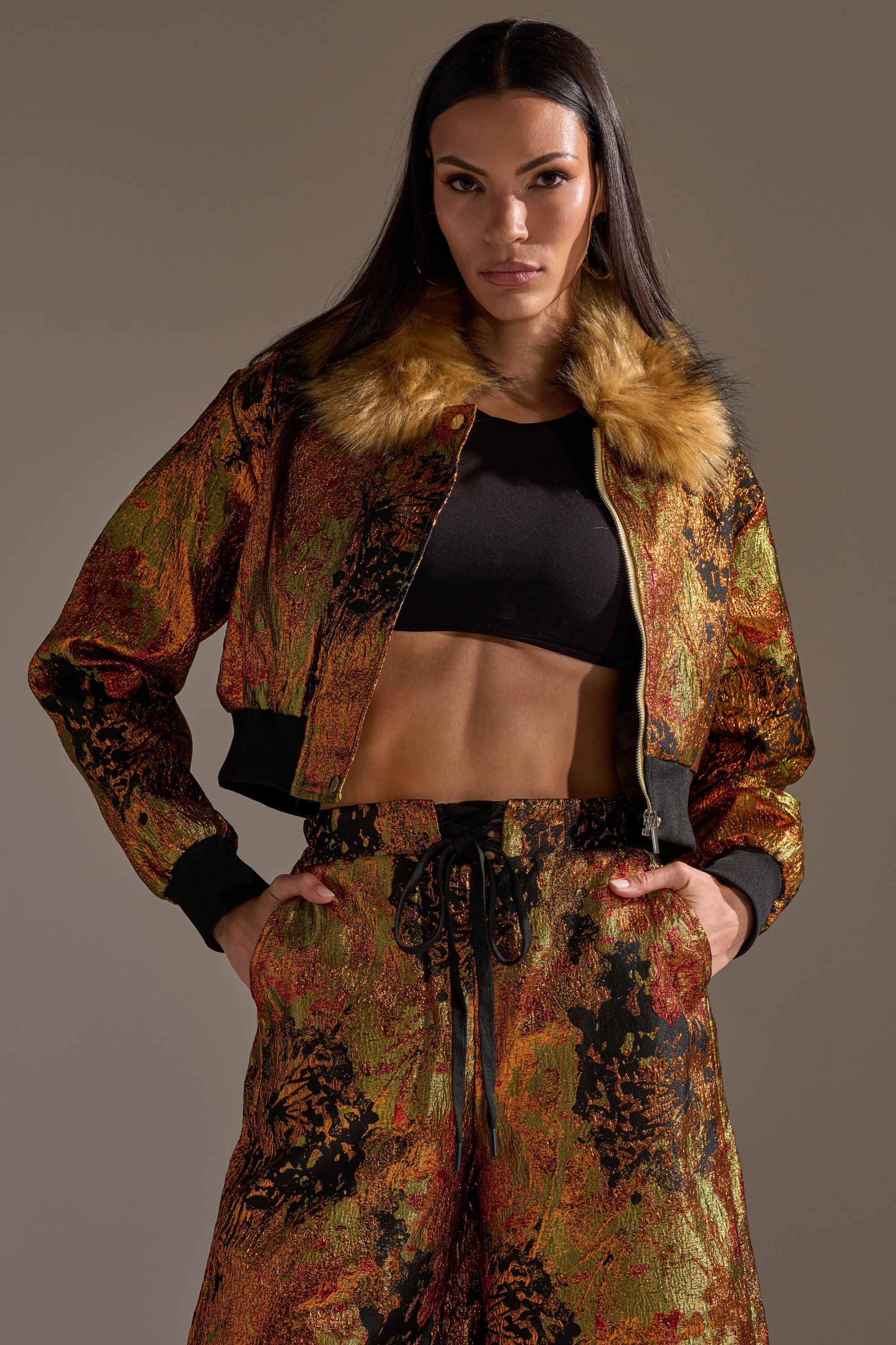 Wearing the TWIN FLAME BROCADE BOMBER with faux fur collar, a woman pairs it with a black crop top and matching multicolored textured pants, standing hands-in-pockets against a neutral backdrop.