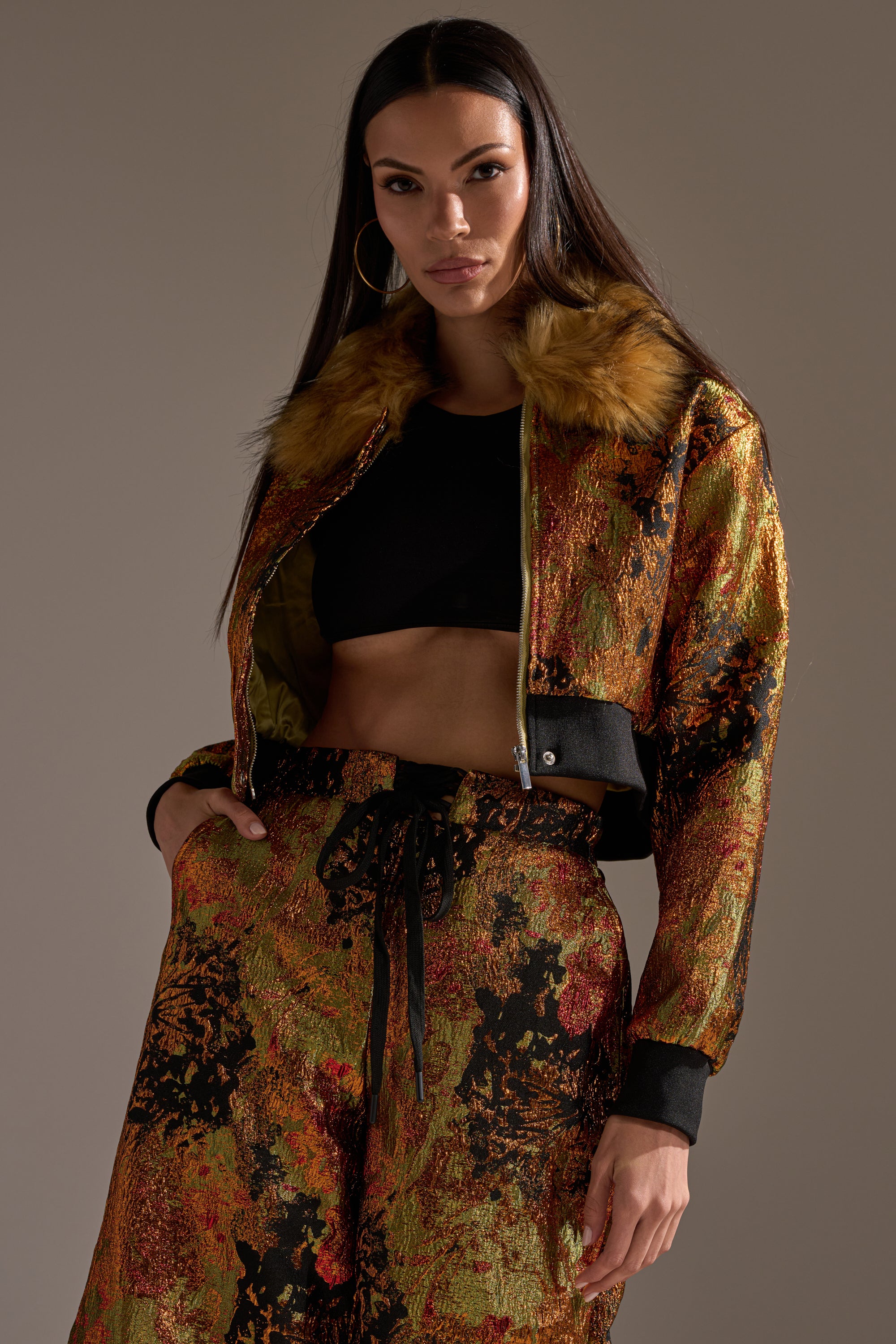 A woman with long dark hair wears the TWIN FLAME BROCADE BOMBER, a black cropped top, and matching gold and red abstract patterned pants with a faux fur collar. She stands confidently against a neutral background.