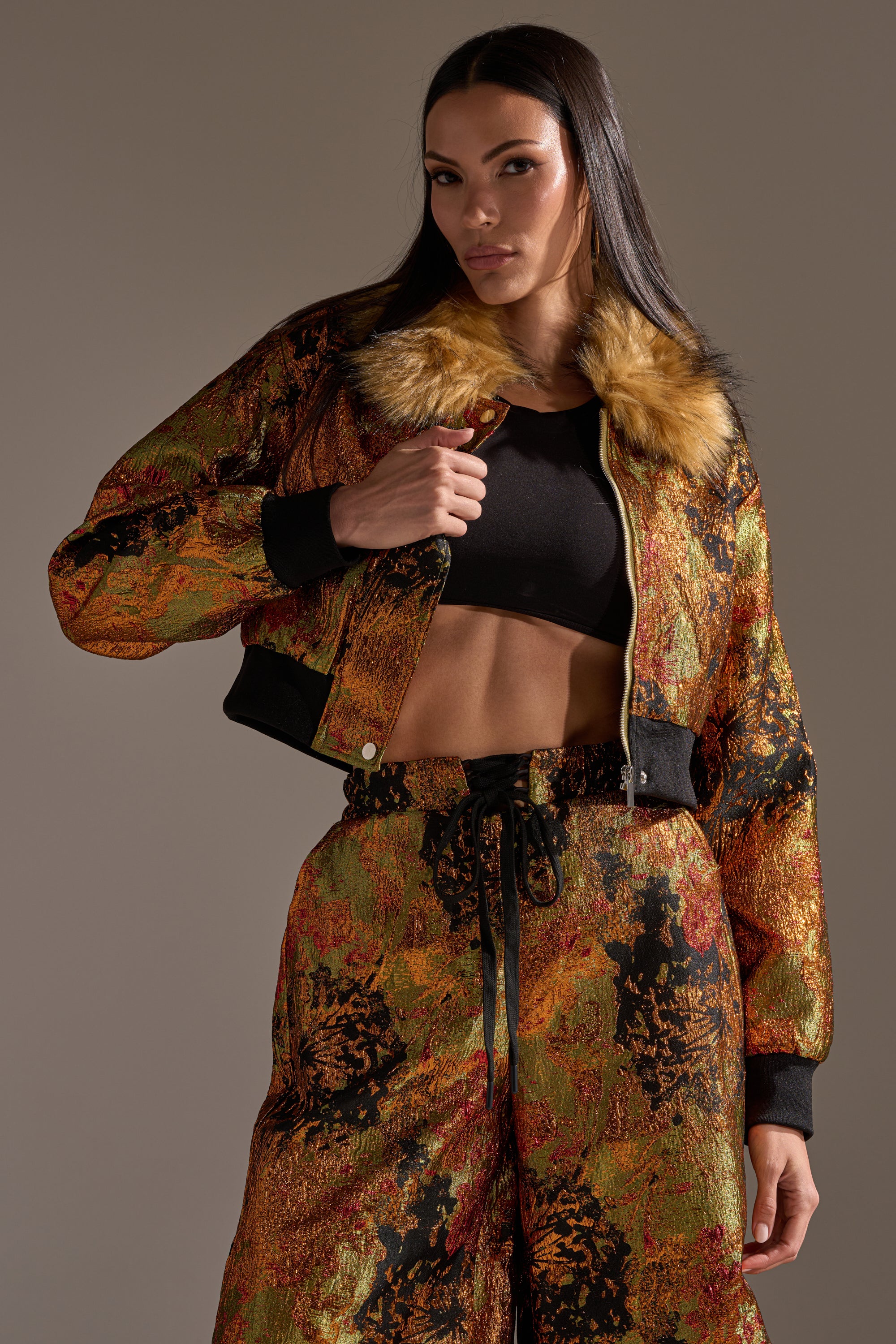 A woman wearing the TWIN FLAME BROCADE BOMBER with a faux fur collar and matching pants in autumnal hues, styled with a black crop top, poses confidently against a neutral background.
