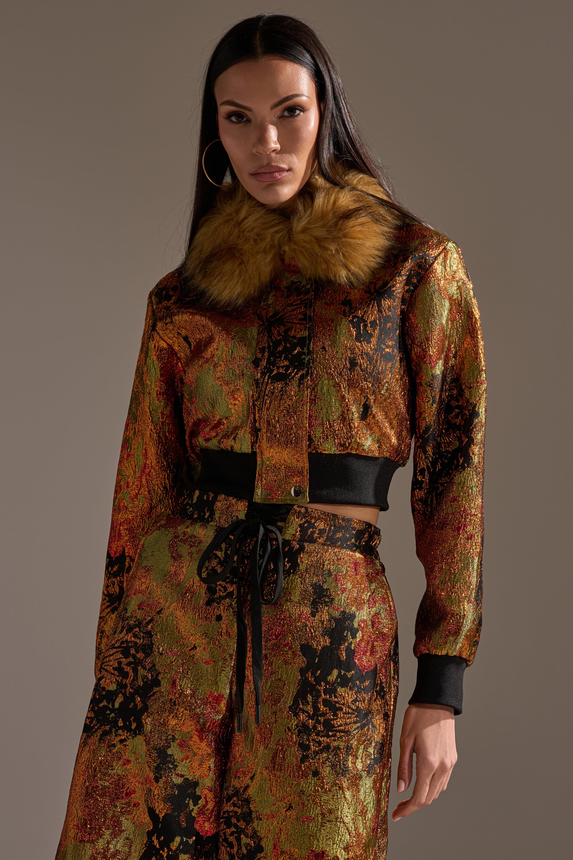 A woman wears the TWIN FLAME BROCADE BOMBER, a cropped metallic brocade jacket with a brown faux fur collar, paired with matching high-waisted pants and a black ribbon belt, standing against a neutral background.