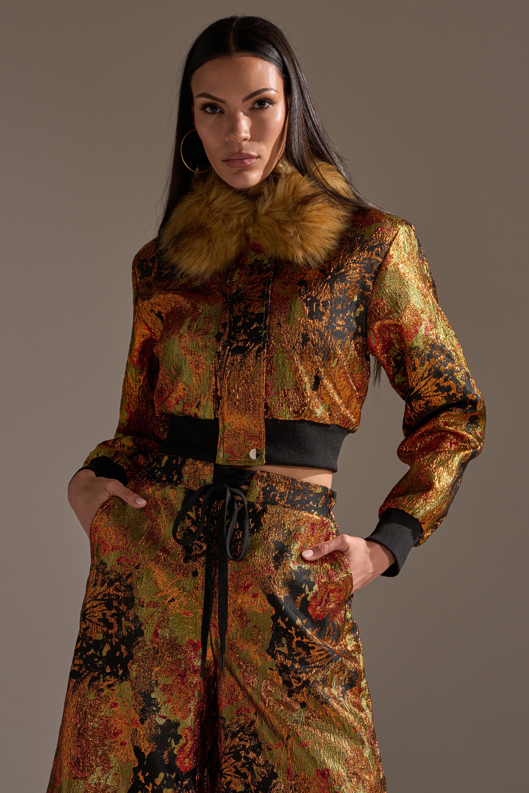 A woman stands confidently with her hands in her pockets, modeling the TWIN FLAME BROCADE BOMBER featuring a metallic brocade finish, faux fur collar, and matching high-waisted pants.
