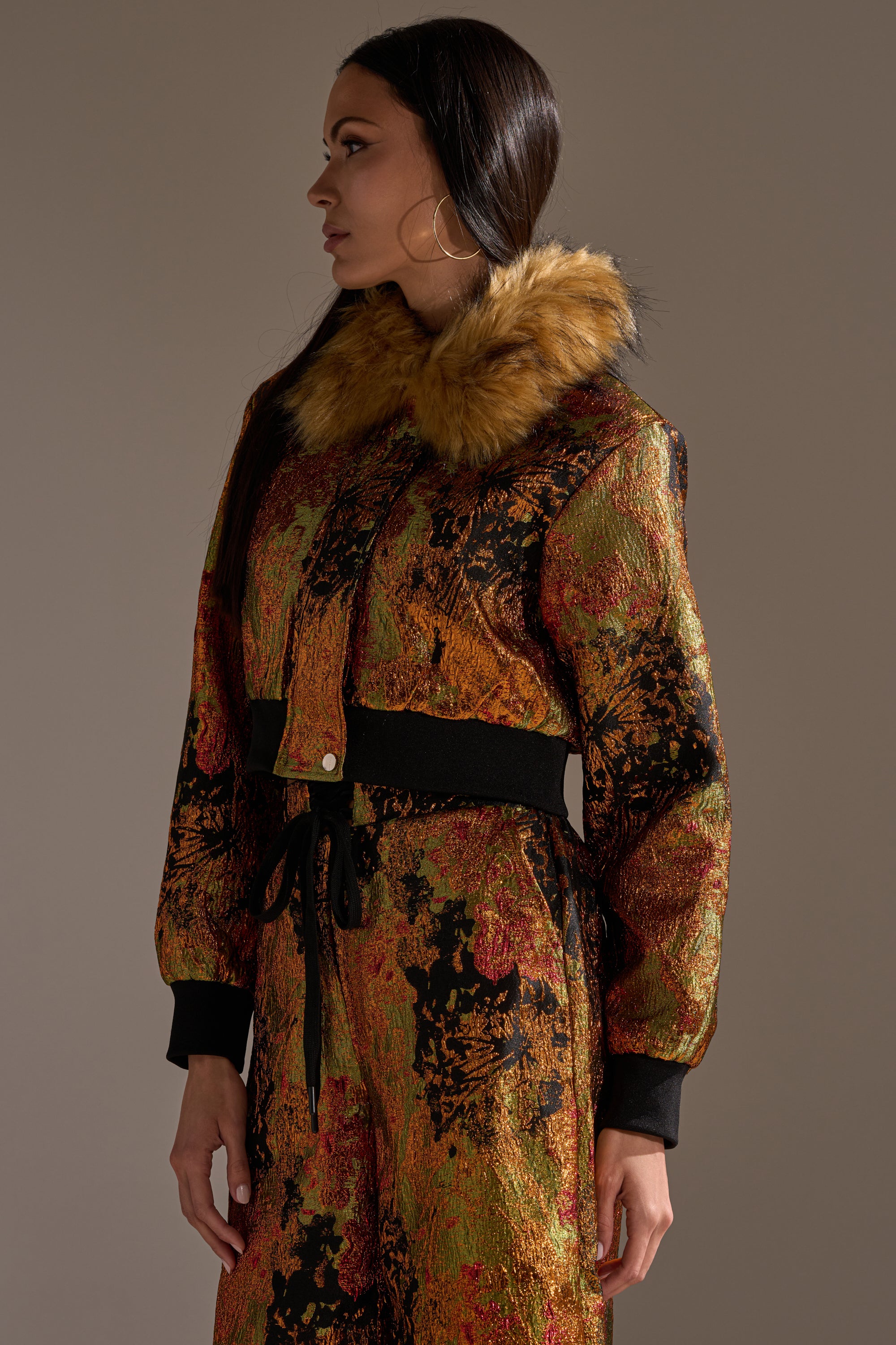 A woman with long dark hair wears the TWIN FLAME BROCADE BOMBER, a cropped, multi-colored metallic jacket with a faux fur collar and black trim, standing against a plain background and looking to her left.