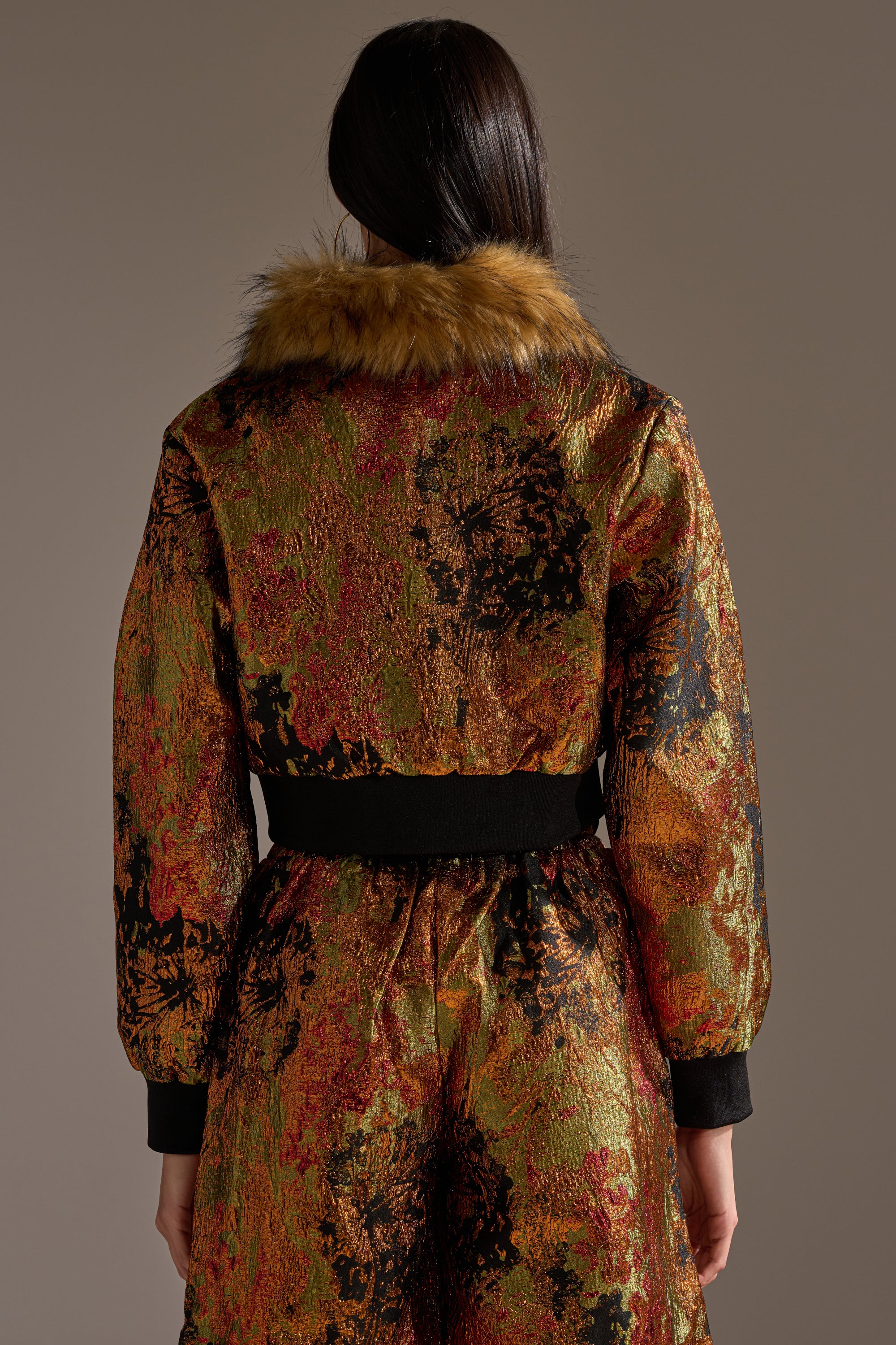 A person with long dark hair is seen from the back wearing the TWIN FLAME BROCADE BOMBER, a cropped jacket with gold, orange, and black abstract patterns, a brown faux fur collar, and black cuffs for standout style.