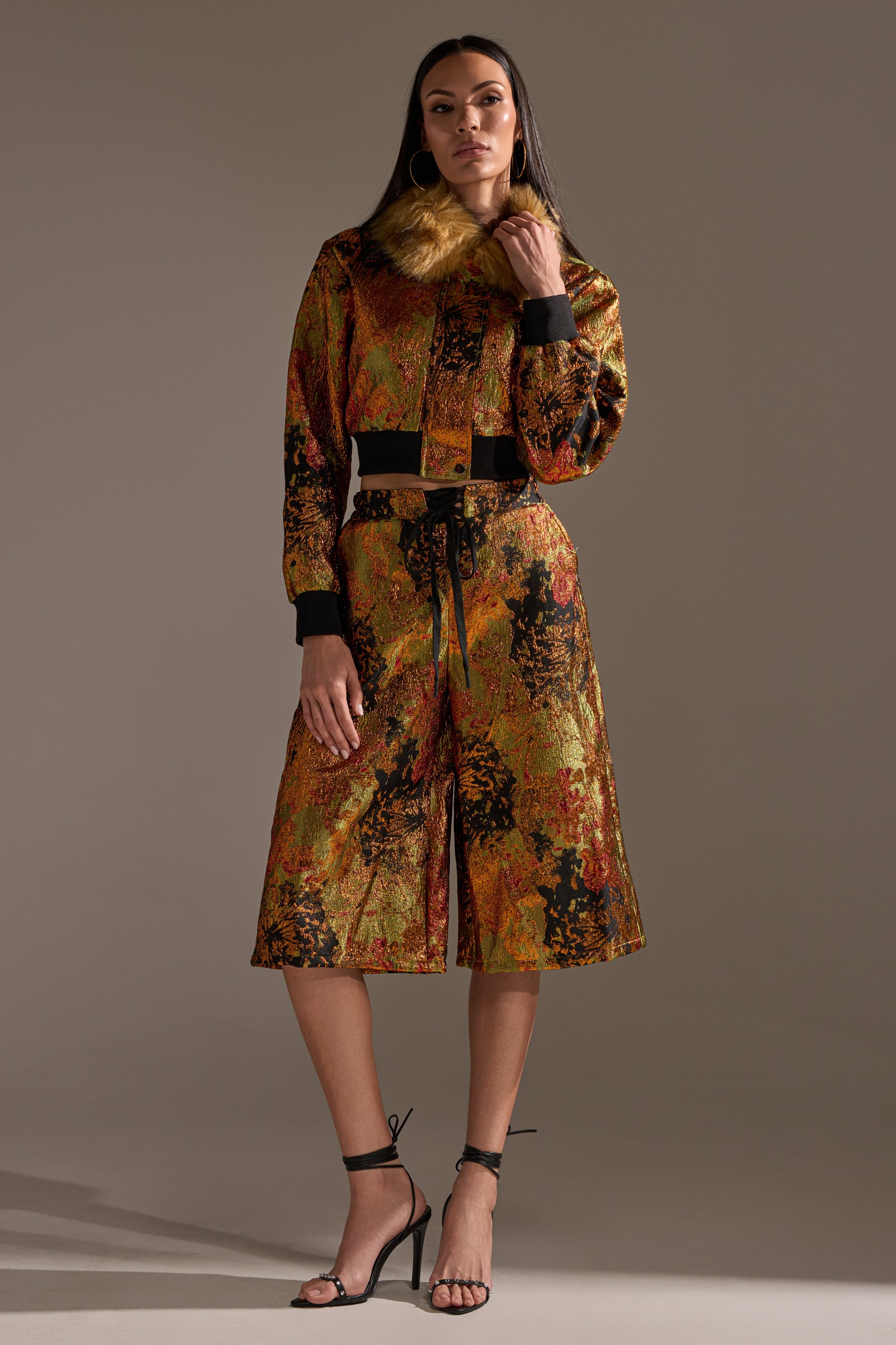 A woman poses confidently against a plain background, wearing the TWIN FLAME BROCADE BOMBER with a faux fur collar and matching culotte pants. She completes the look with long dark hair and black high-heeled sandals.