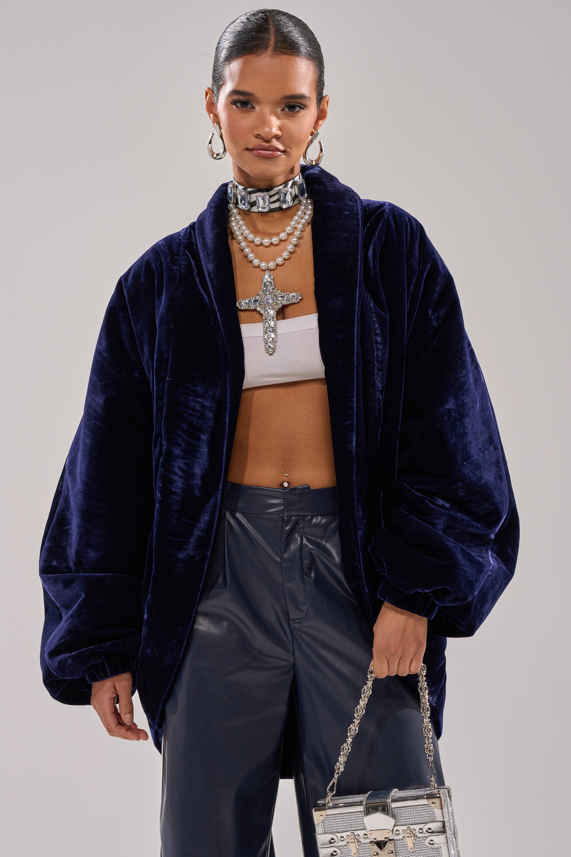 A woman wearing the LOLA COCOON COAT IN VELVET (royal blue), navy leather pants, a white crop top, layered pearl necklaces with a jeweled cross, hoop earrings, and carrying a metallic silver handbag.