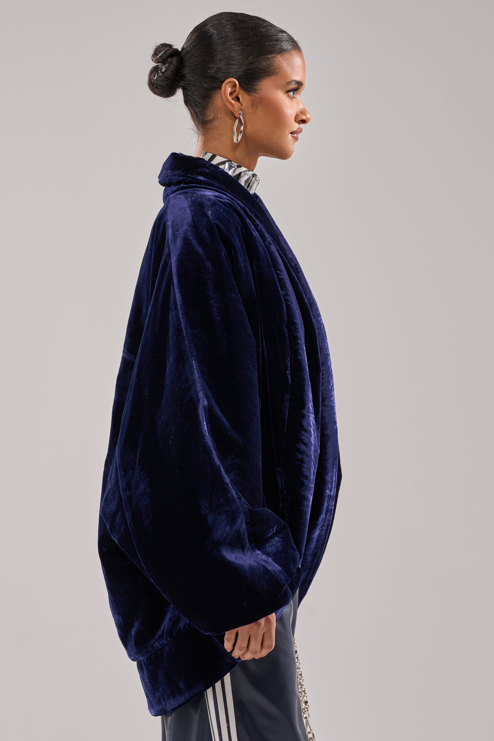 A woman with dark hair in a bun models the LOLA COCOON COAT IN VELVET, paired with a patterned turtleneck and hoop earrings, standing in profile against a plain light background.