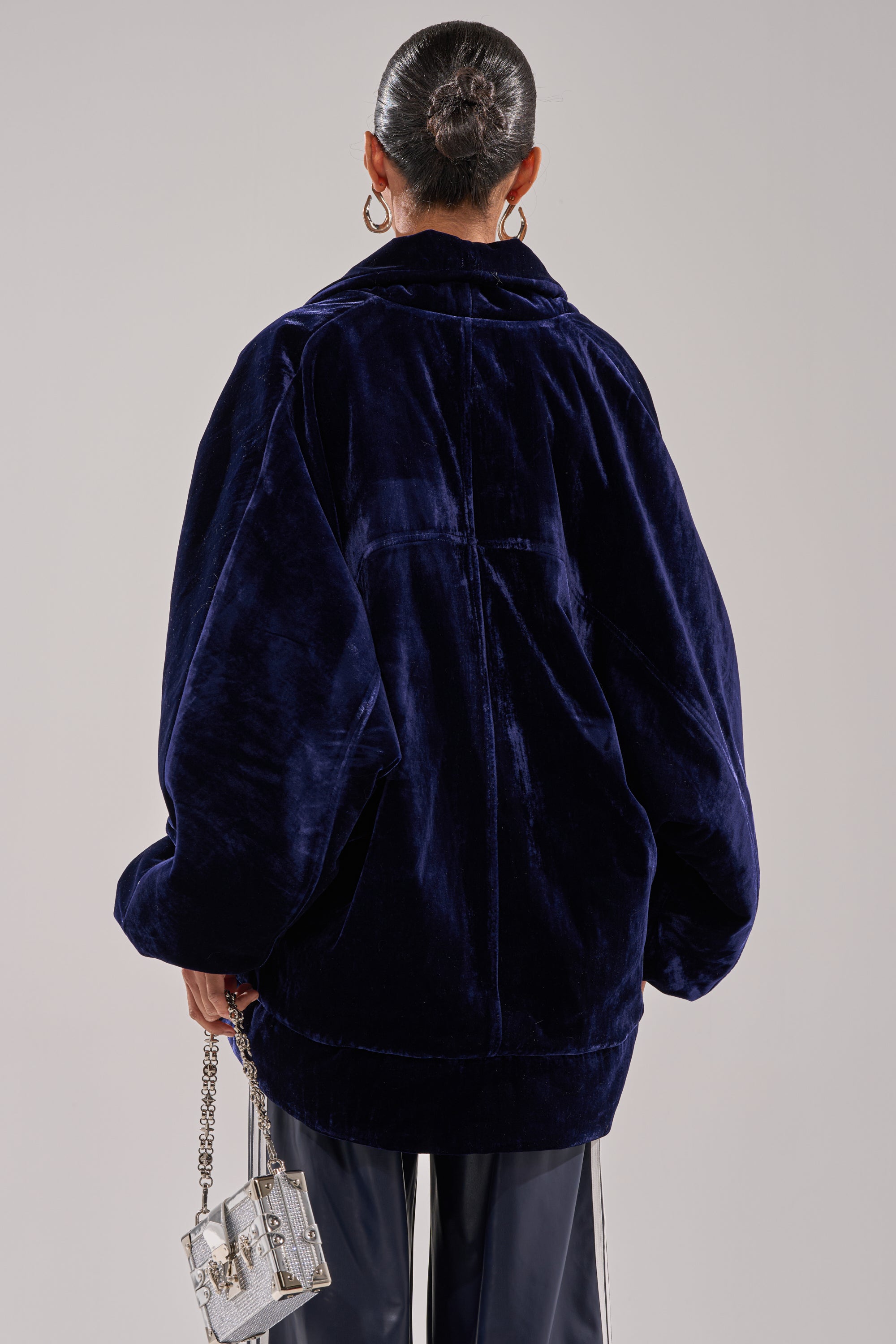 With her hair in a sleek bun, a person faces away wearing the LOLA COCOON COAT IN VELVET in dark blue, styled with black pants and a small clear handbag with a silver chain, set against a plain background.