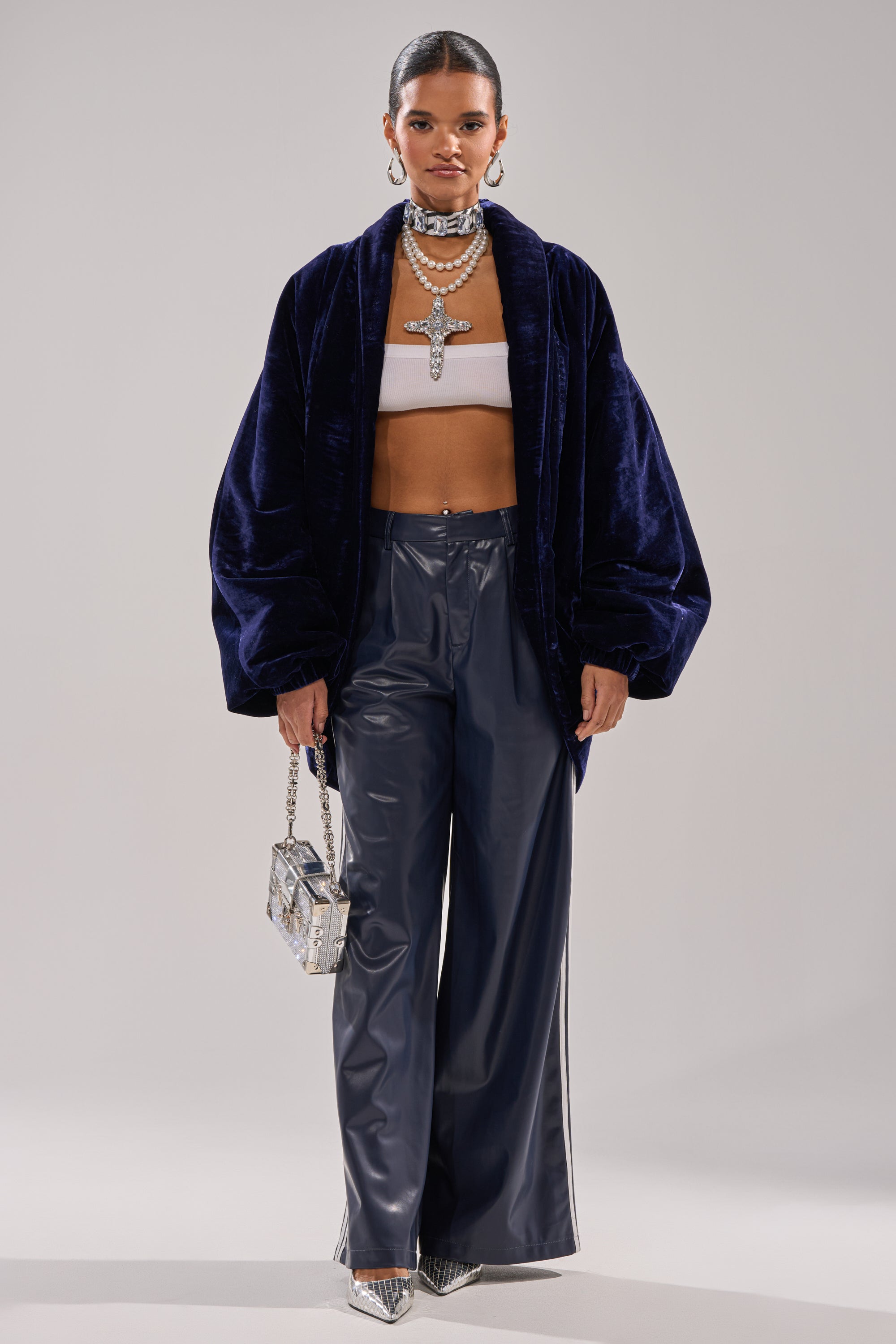 A woman models the LOLA COCOON COAT IN VELVET, paired with a white bandeau, wide-leg navy pants, silver pointed heels, bold layered jewelry, and a small metallic purse against a plain background.
