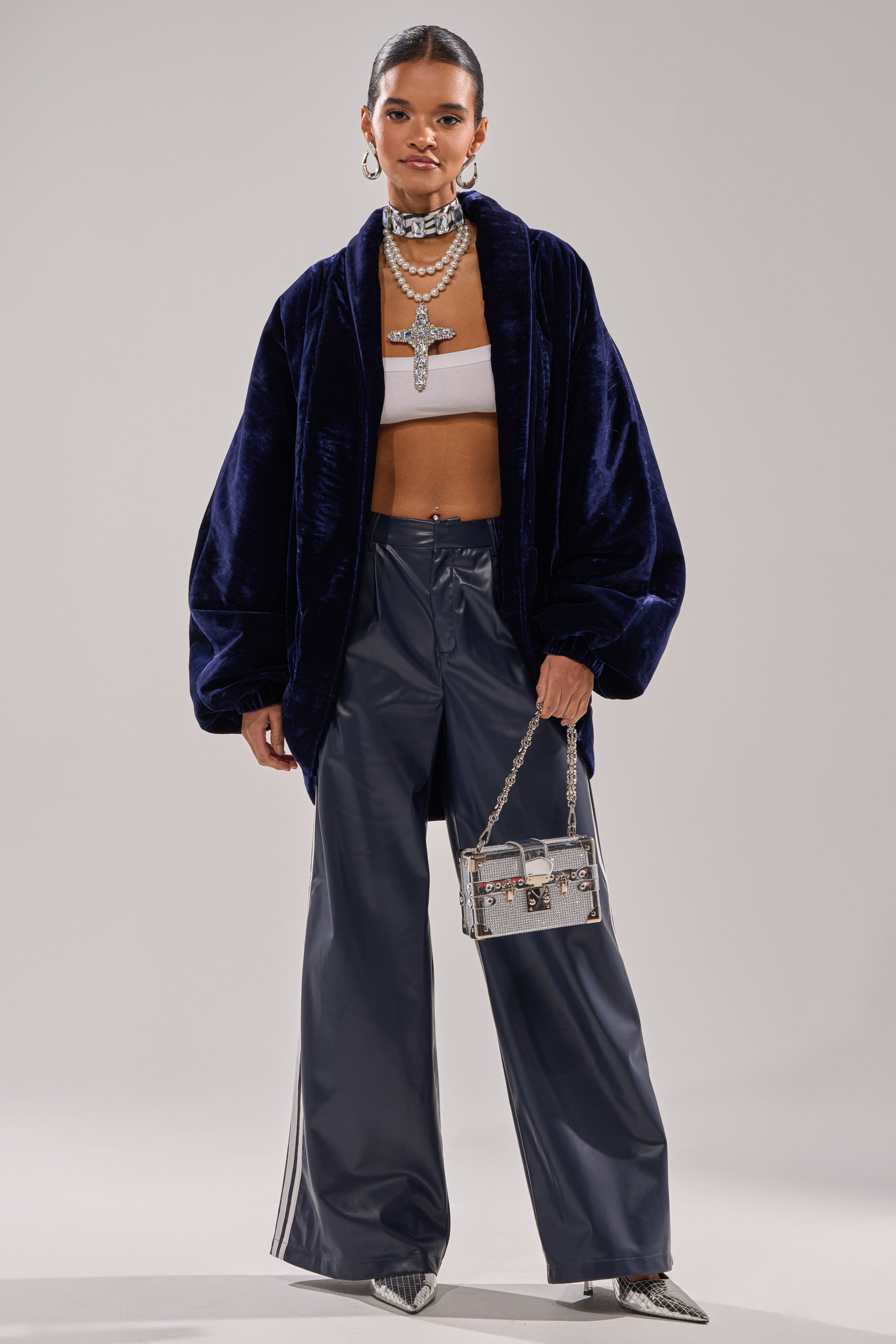 A woman stands confidently in the LOLA COCOON COAT IN VELVET, paired with a white bandeau top, wide-leg shiny navy pants, pointed silver heels, layered silver necklaces, and a small silver handbag.