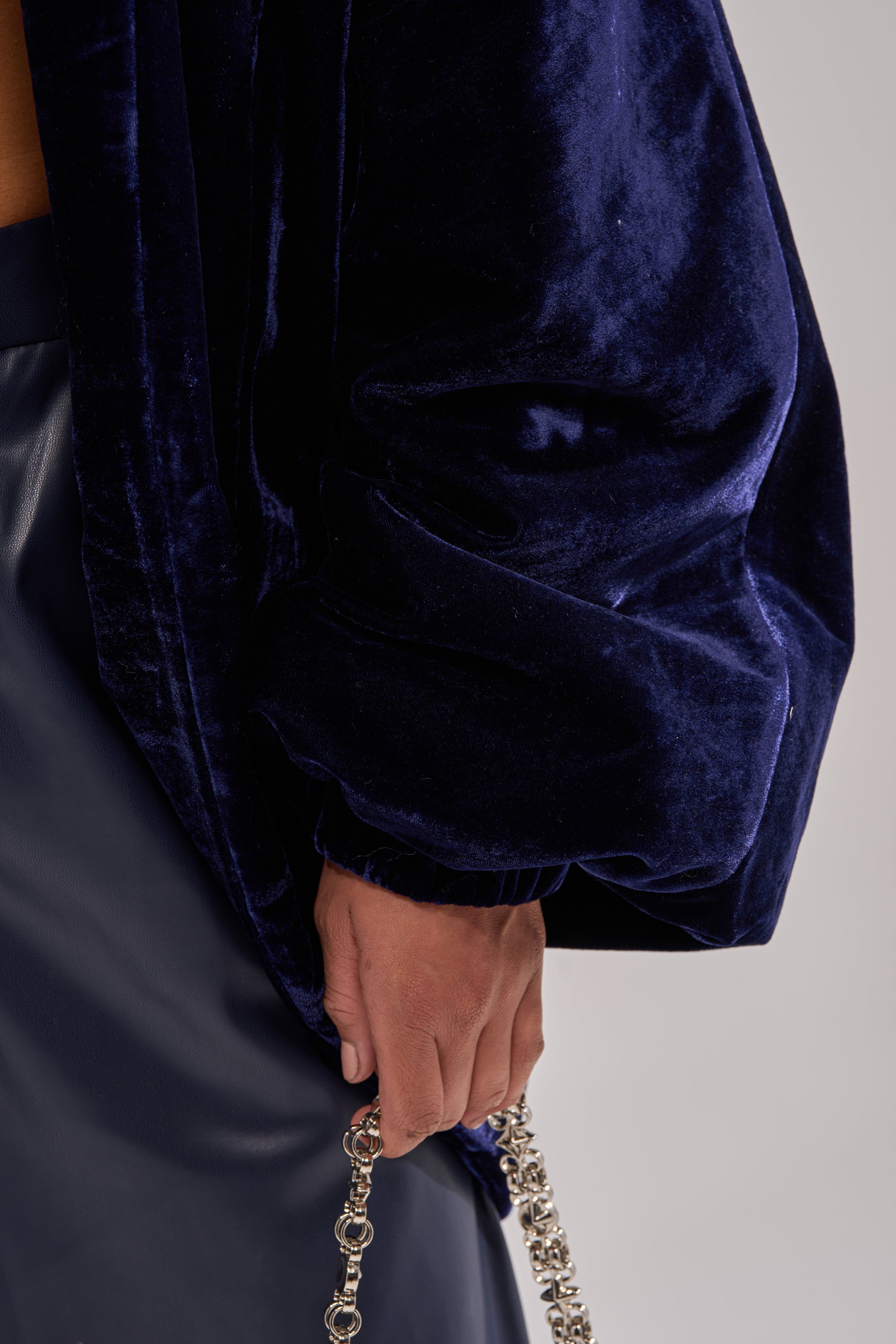 A person wears the LOLA COCOON COAT IN VELVET and holds a silver chain, while the background remains out of focus.