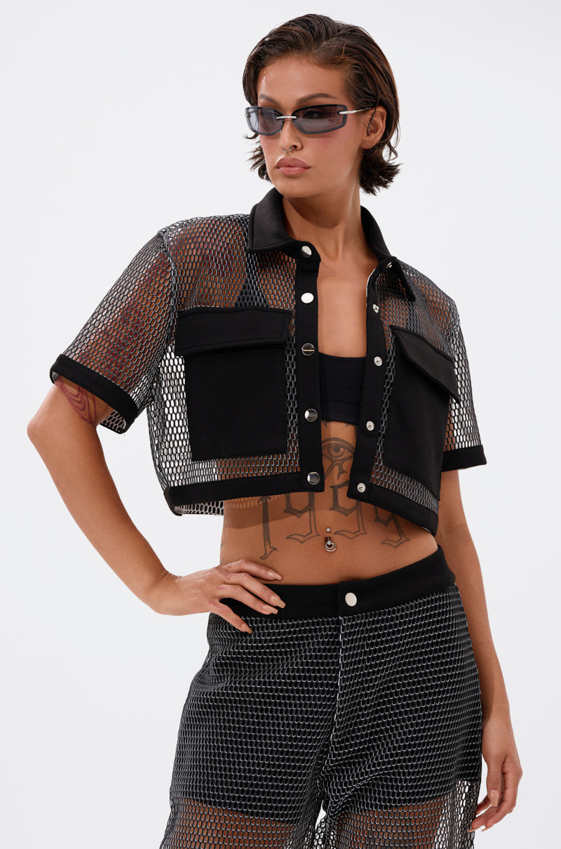 A person with short hair and visible torso tattoos wears the STEPHANIA TRANSPARENT BUTTON DOWN—a black fishnet mesh, cropped shirt with pocket details—paired with matching mesh shorts. They wear sunglasses against a plain white background.