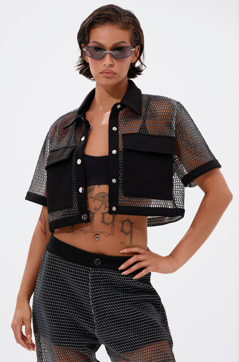 A woman poses confidently against a plain background wearing sunglasses, the STEPHANIA TRANSPARENT BUTTON DOWN with black panels and buttons over a black bandeau, and matching mesh shorts.