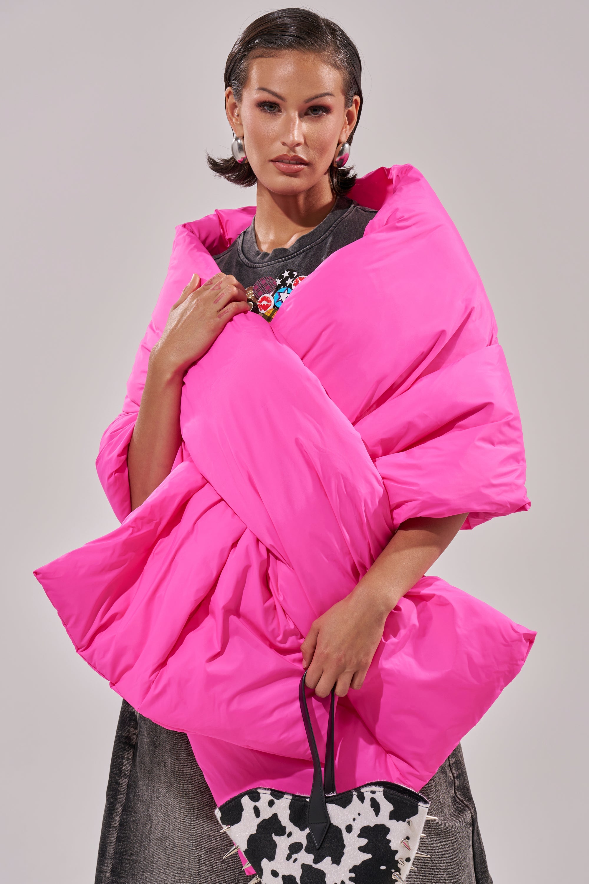 A woman with short dark hair models the WRAPPED UP PUFFER SCARF IN PINK over a dark top and grey skirt. She confidently holds a black-and-white cow print handbag, posing against a plain background. 0