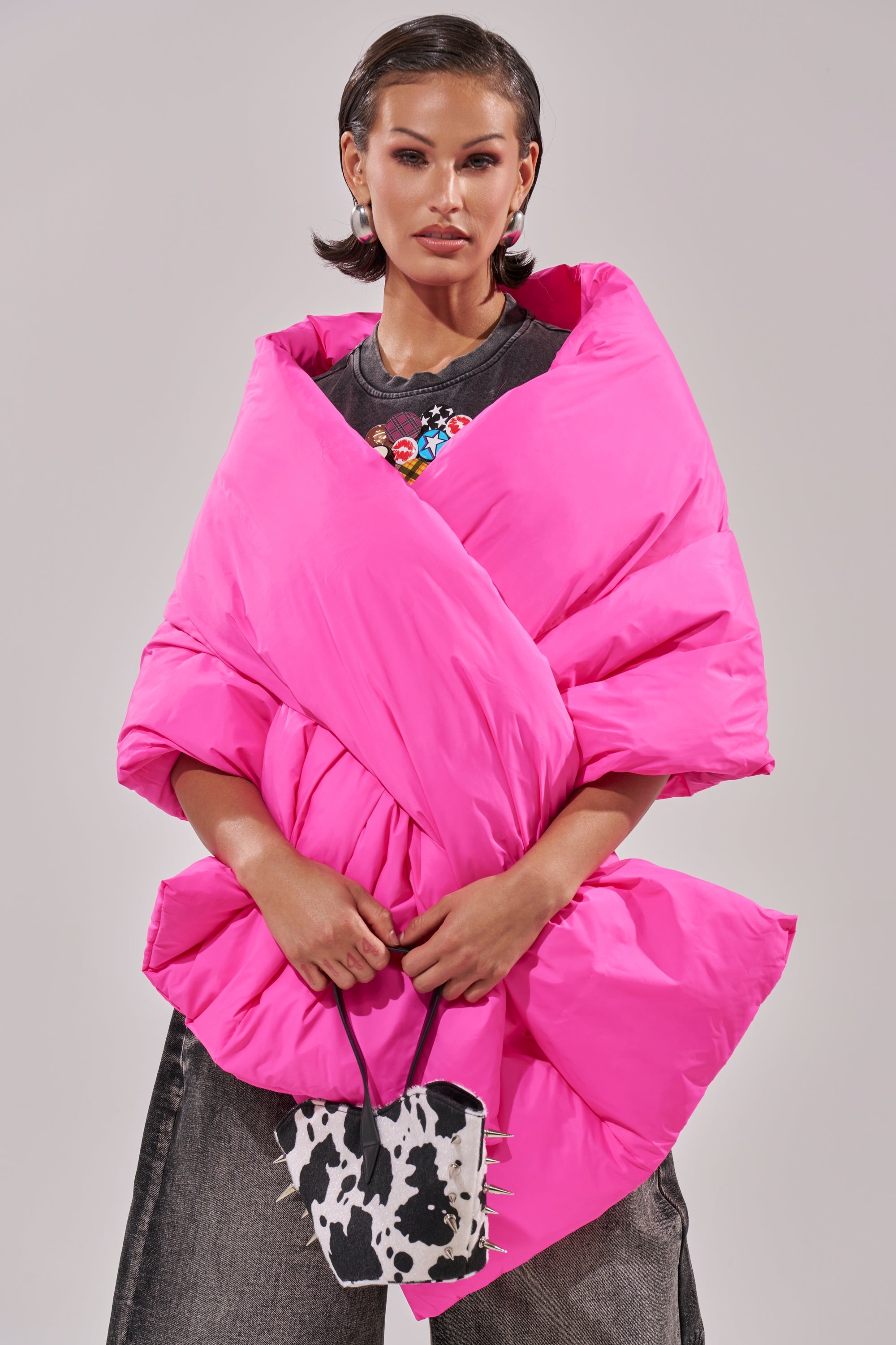 A woman stands confidently against a light background in dark wide-leg jeans, a bright pink oversized puffer jacket, and a small cow-print handbag—her look finished with the WRAPPED UP PUFFER SCARF IN PINK. 1