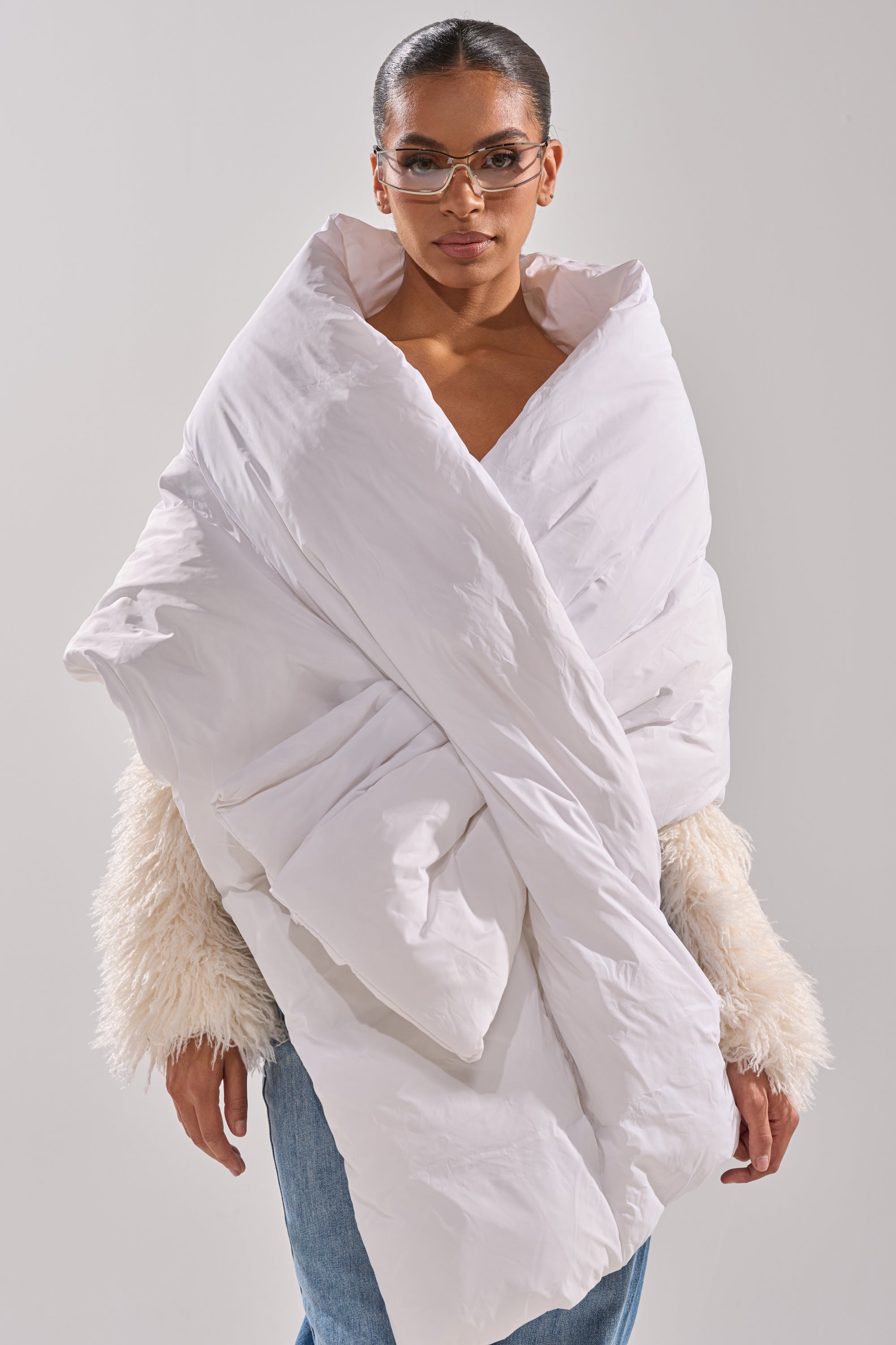 A person stands against a plain light background wearing large, stylish glasses, a faux fur sweater, blue jeans, and the WRAPPED UP PUFFER SCARF IN WHITE. 0