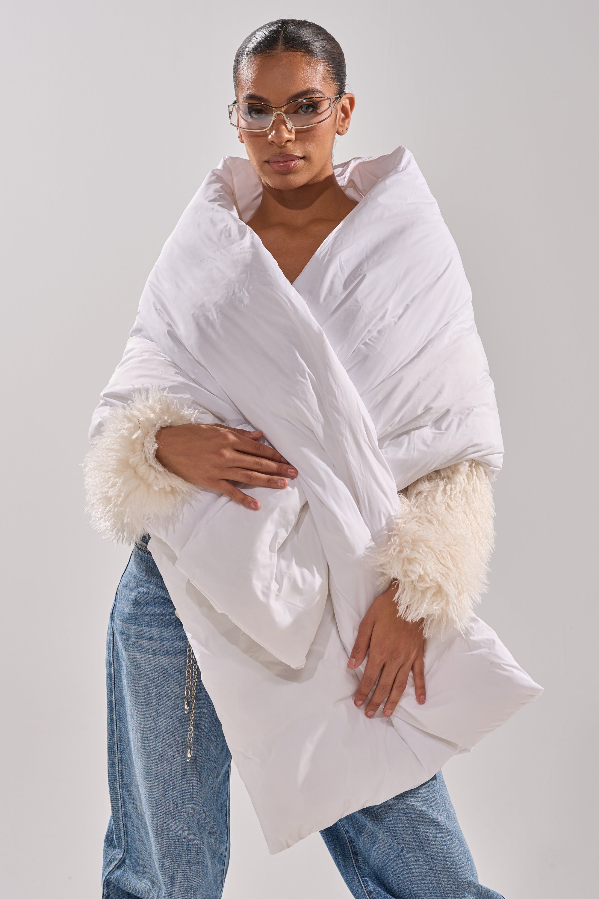 A woman poses confidently against a plain, light background, wearing sunglasses, a white fluffy-sleeved top, blue jeans, and the WRAPPED UP PUFFER SCARF IN WHITE draped over her shoulders. 1
