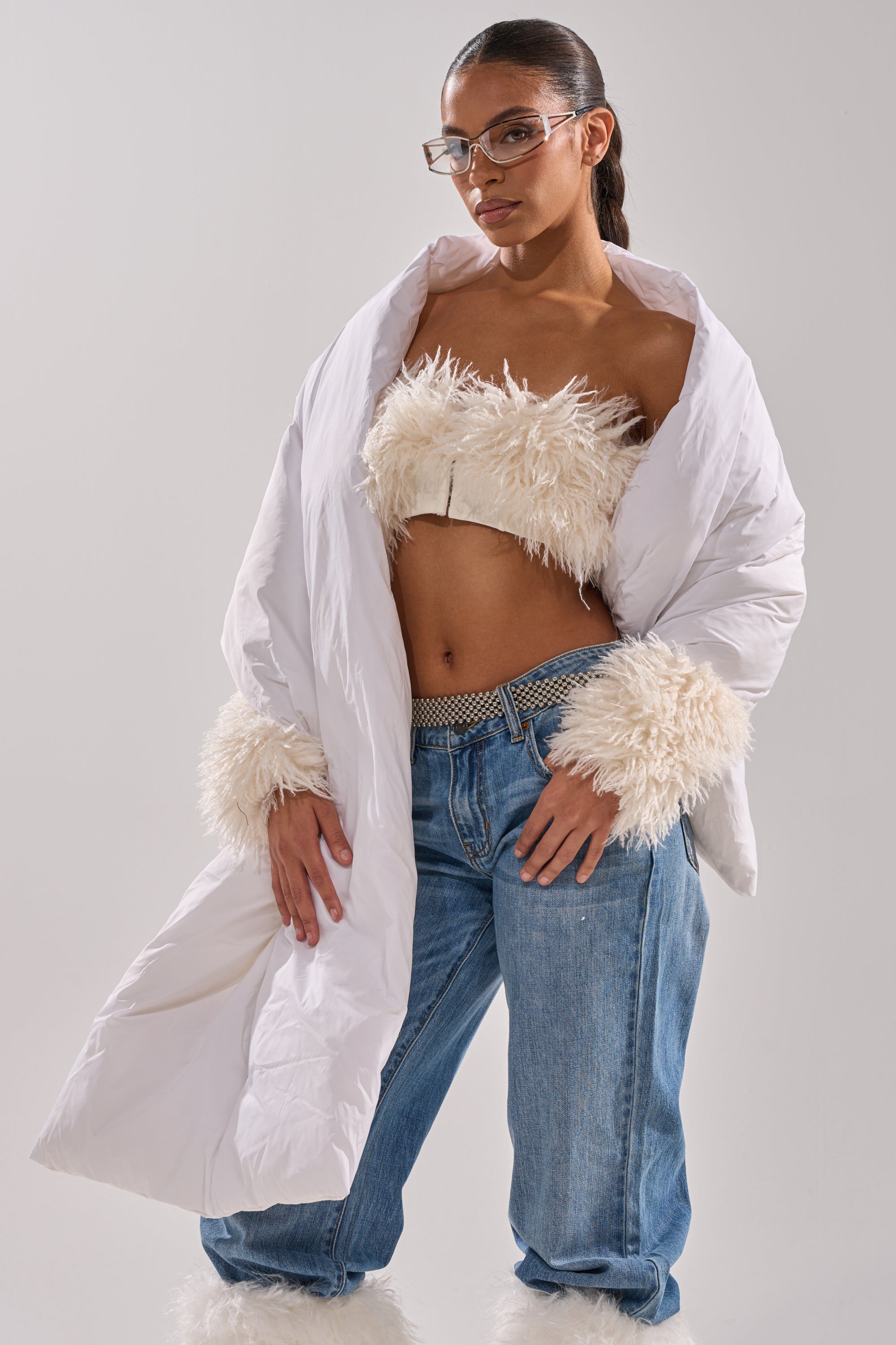 A woman poses confidently in a white fluffy crop top, a white puffy coat with furry cuffs, loose blue jeans, glasses, and her hair tied back, accessorized with the WRAPPED UP PUFFER SCARF IN WHITE and a studded belt. 2