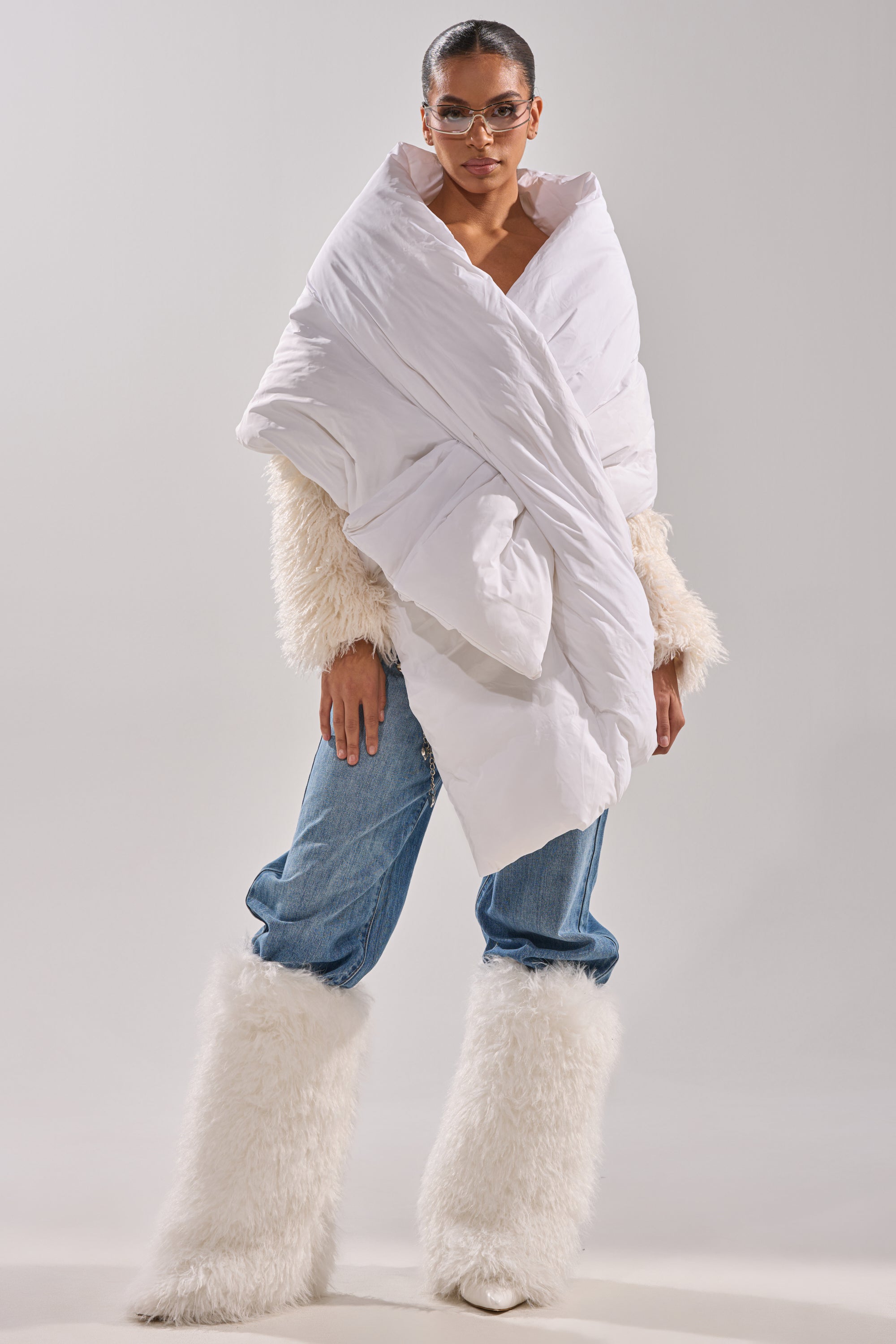 A person stands confidently against a plain light background, wearing the WRAPPED UP PUFFER SCARF IN WHITE with fluffy cream sleeves, blue jeans, and tall furry white boots. 3