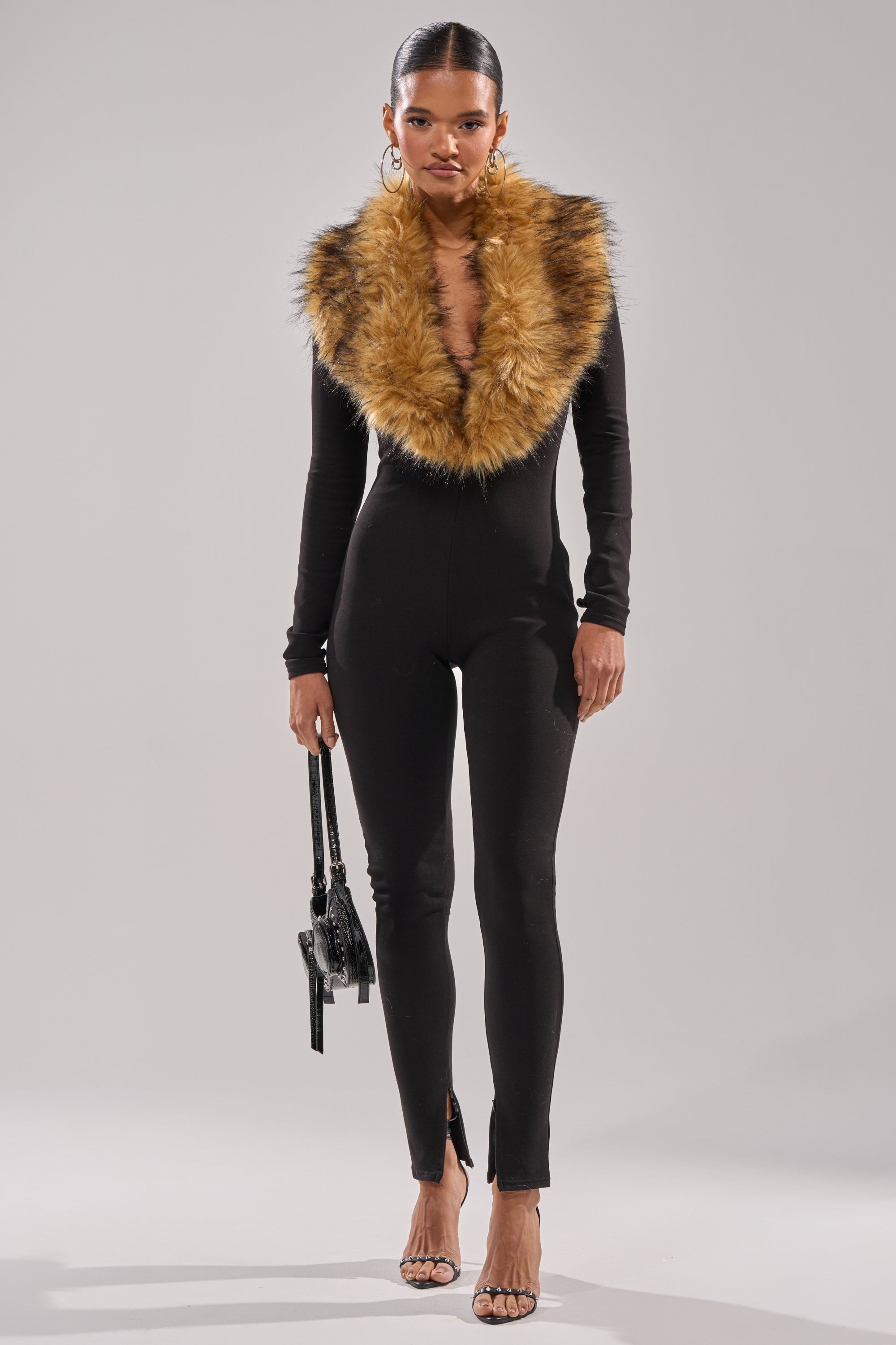 A woman faces forward in the OH HOT DAMN BABY FUR TRIM JUMPSUIT featuring a fitted silhouette and deep V neckline, accessorized with a black handbag and strappy heels—an ideal look for a luxurious night out.