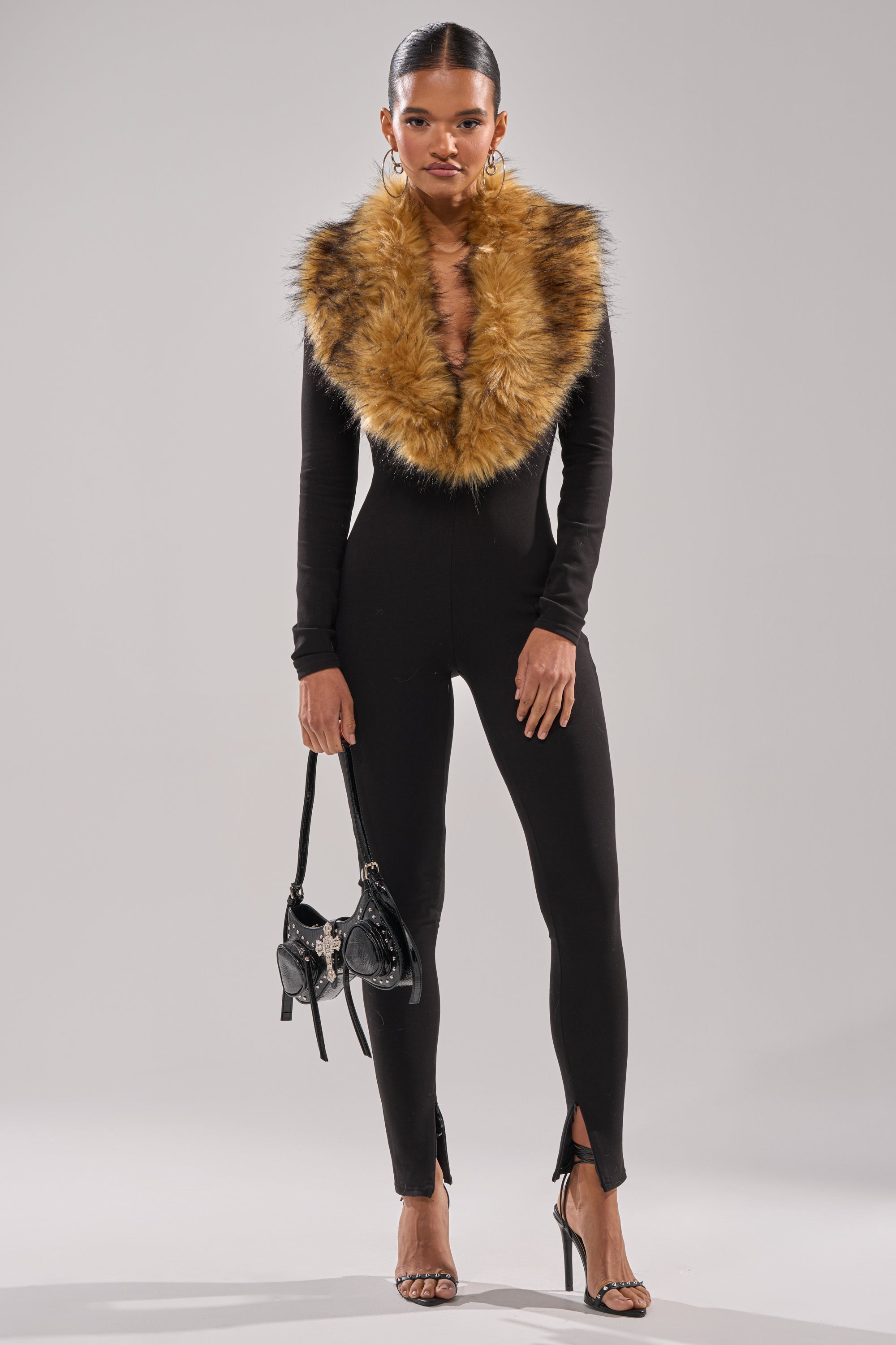 A woman exudes confidence in the OH HOT DAMN BABY FUR TRIM JUMPSUIT, featuring a black design with a deep V-neck and fur trim, styled with a black handbag and heels—perfect for an elegant holiday party look.