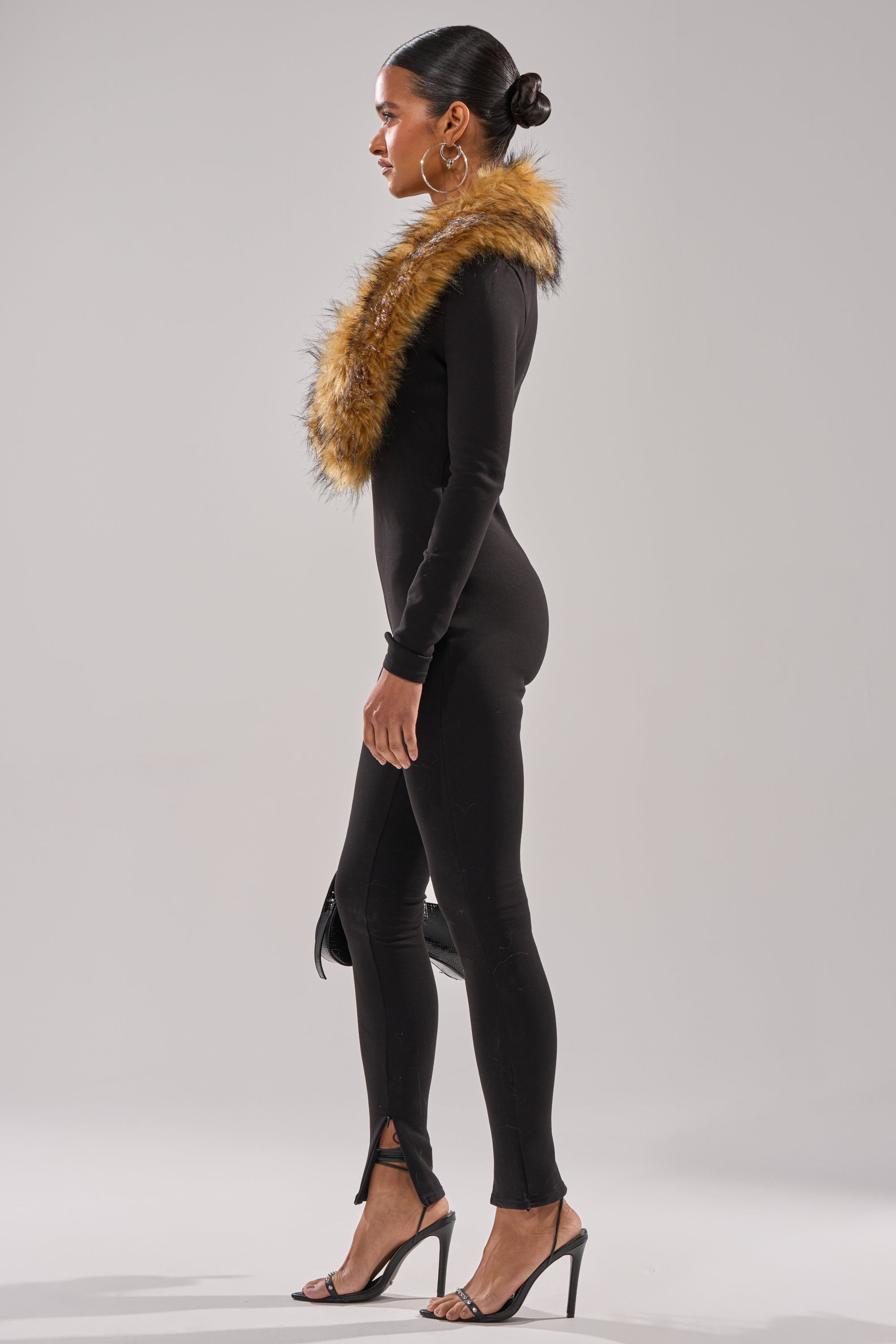 A woman in profile wears the OH HOT DAMN BABY FUR TRIM JUMPSUIT with a brown faux fur stole, black high-heeled sandals, and a black clutch. Her hair is in a sleek bun, creating a chic holiday party look against a plain light background.