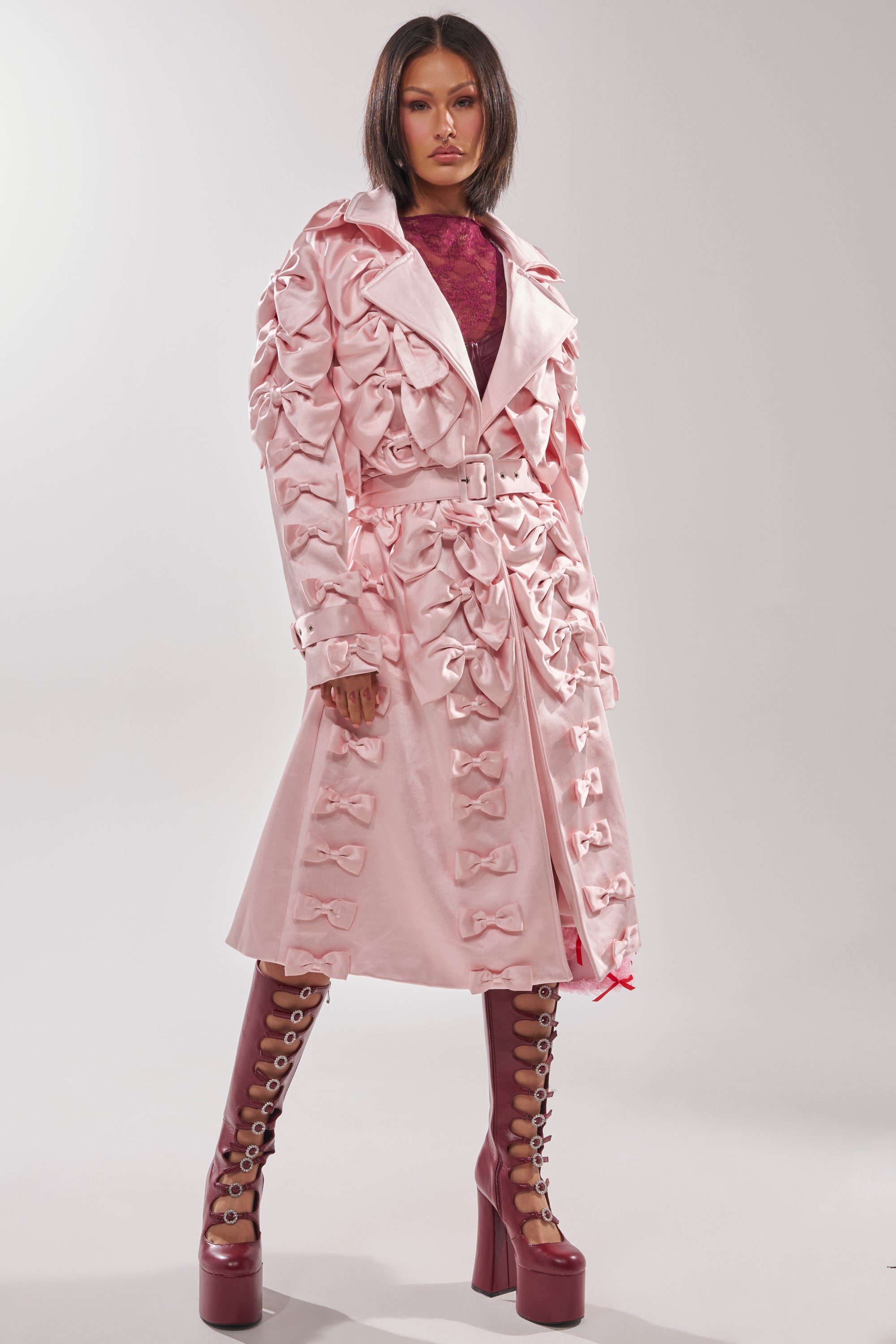 A woman poses confidently against a plain light background, wearing the WRAP ME WITH A SATIN BOW TRENCH—a couture-inspired light pink coat with multiple satin bows—paired with a maroon top and maroon platform lace-up boots.