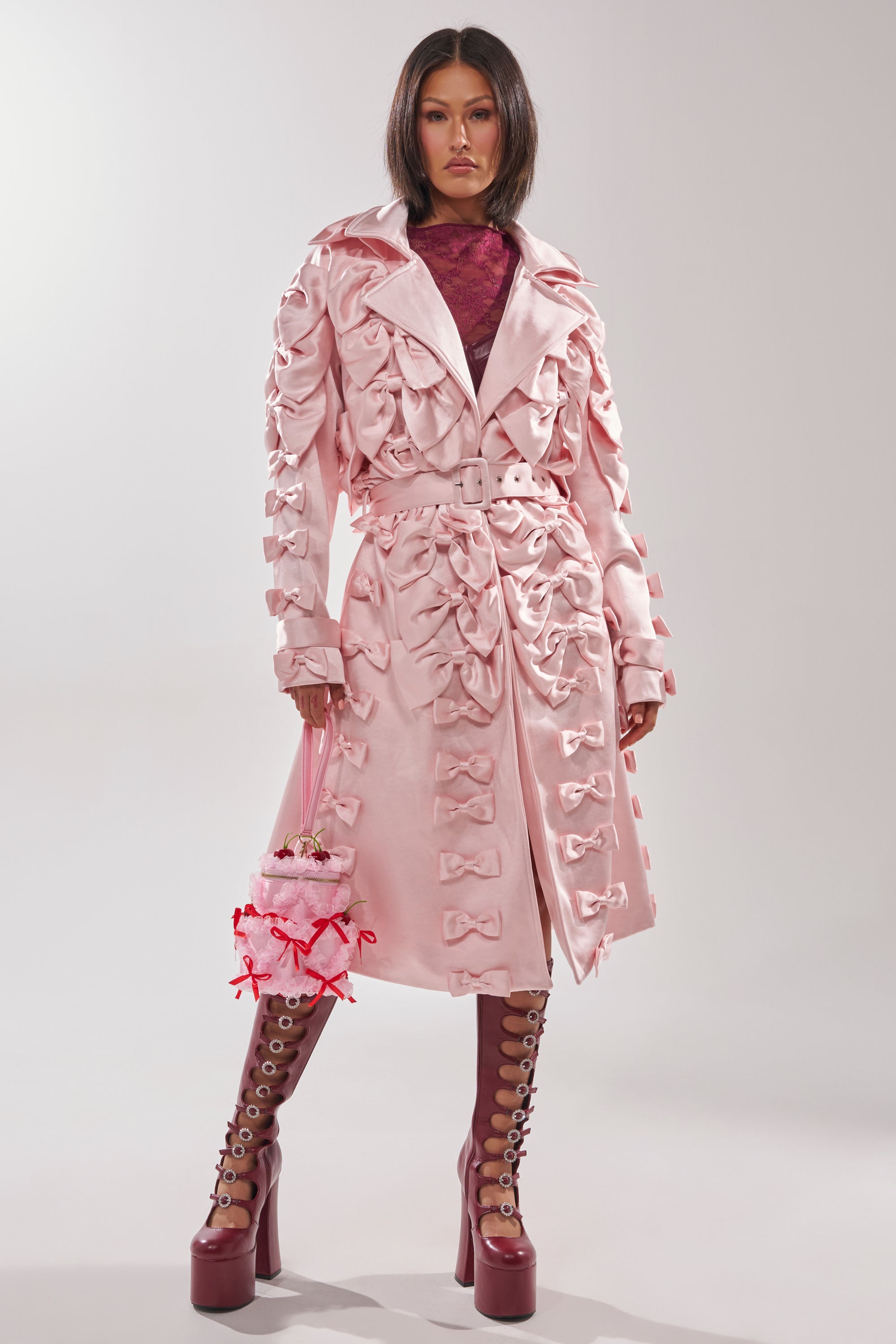 A woman stands confidently in the WRAP ME WITH A SATIN BOW TRENCH, adorned with bows and styled with burgundy lace-up platform boots and a floral handbag. She has straight dark hair and a neutral expression.