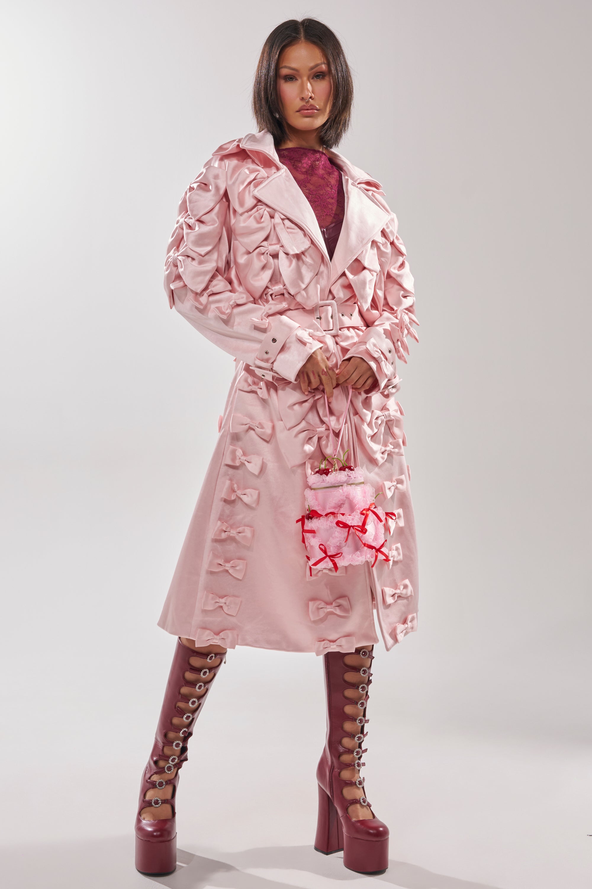 A woman stands confidently in the WRAP ME WITH A SATIN BOW TRENCH, a pale pink, knee-length coat with multiple bows. She carries a small pink handbag and wears maroon lace-up platform boots against a plain light gray background.