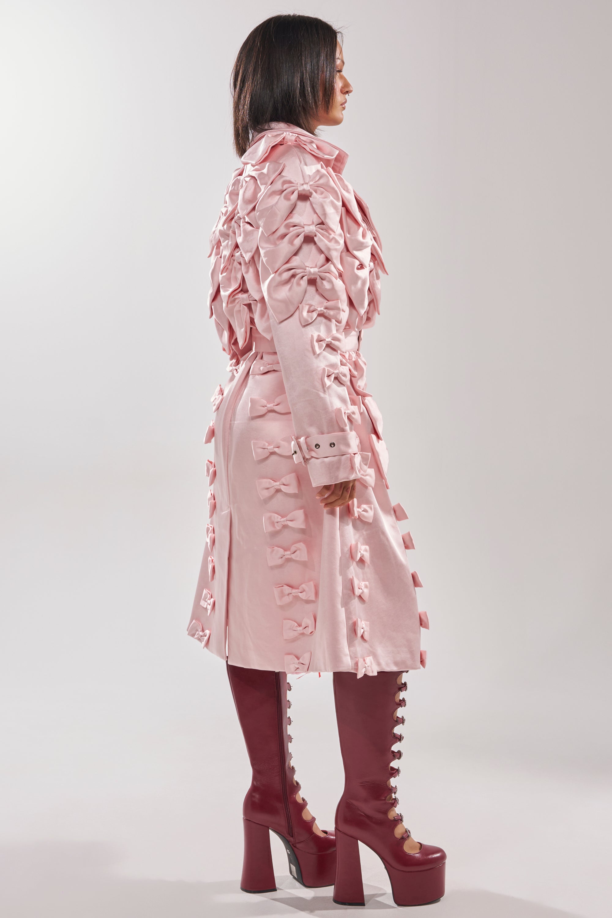 A person stands in profile wearing the WRAP ME WITH A SATIN BOW TRENCH, a light pink coat detailed with multiple bows, paired with knee-high maroon lace-up platform boots against a plain light background.