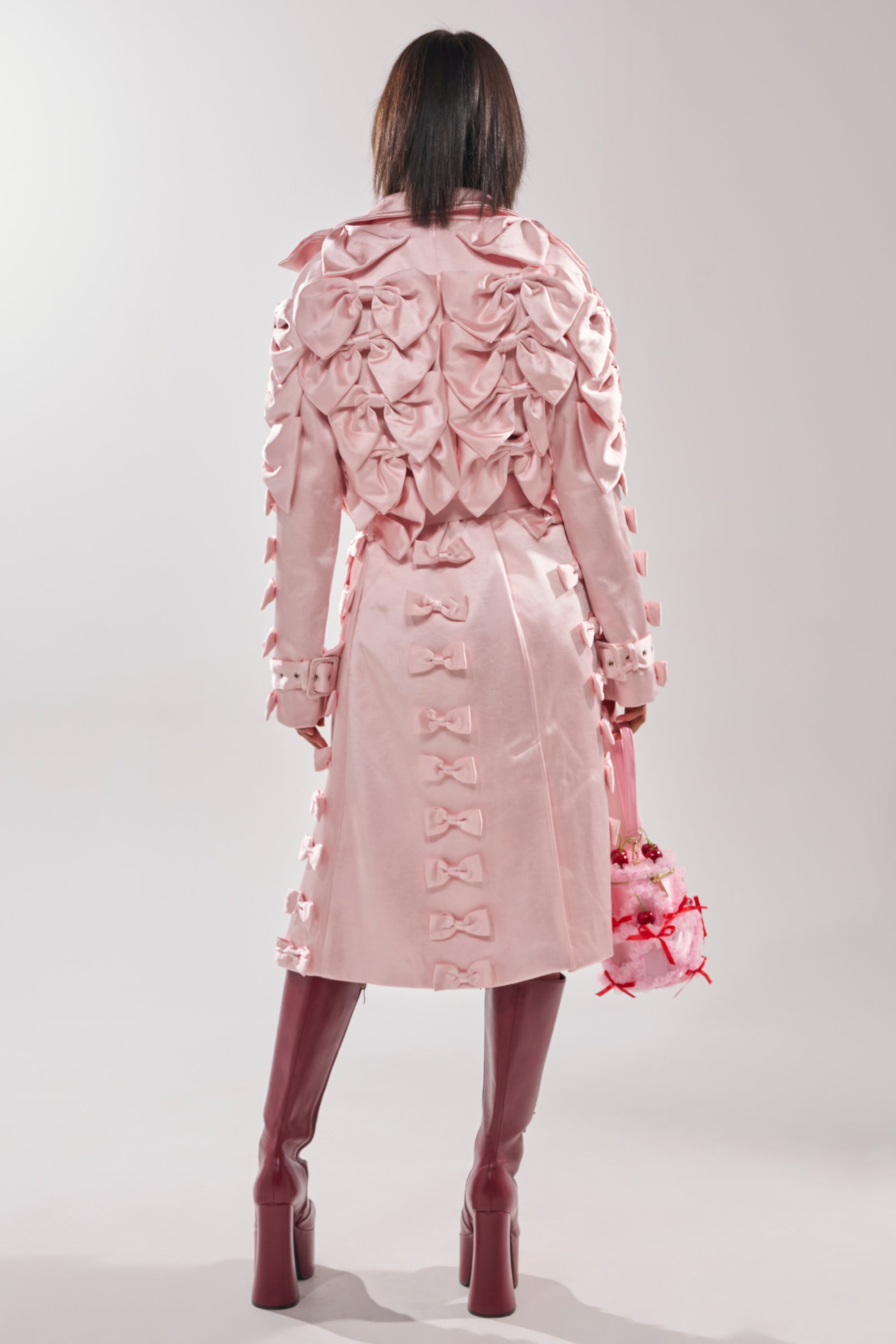 A person faces away in the WRAP ME WITH A SATIN BOW TRENCH, a pale pink, couture-inspired coat adorned with bows. They carry a pink floral bag and wear tall dark mauve platform boots against a plain light background.
