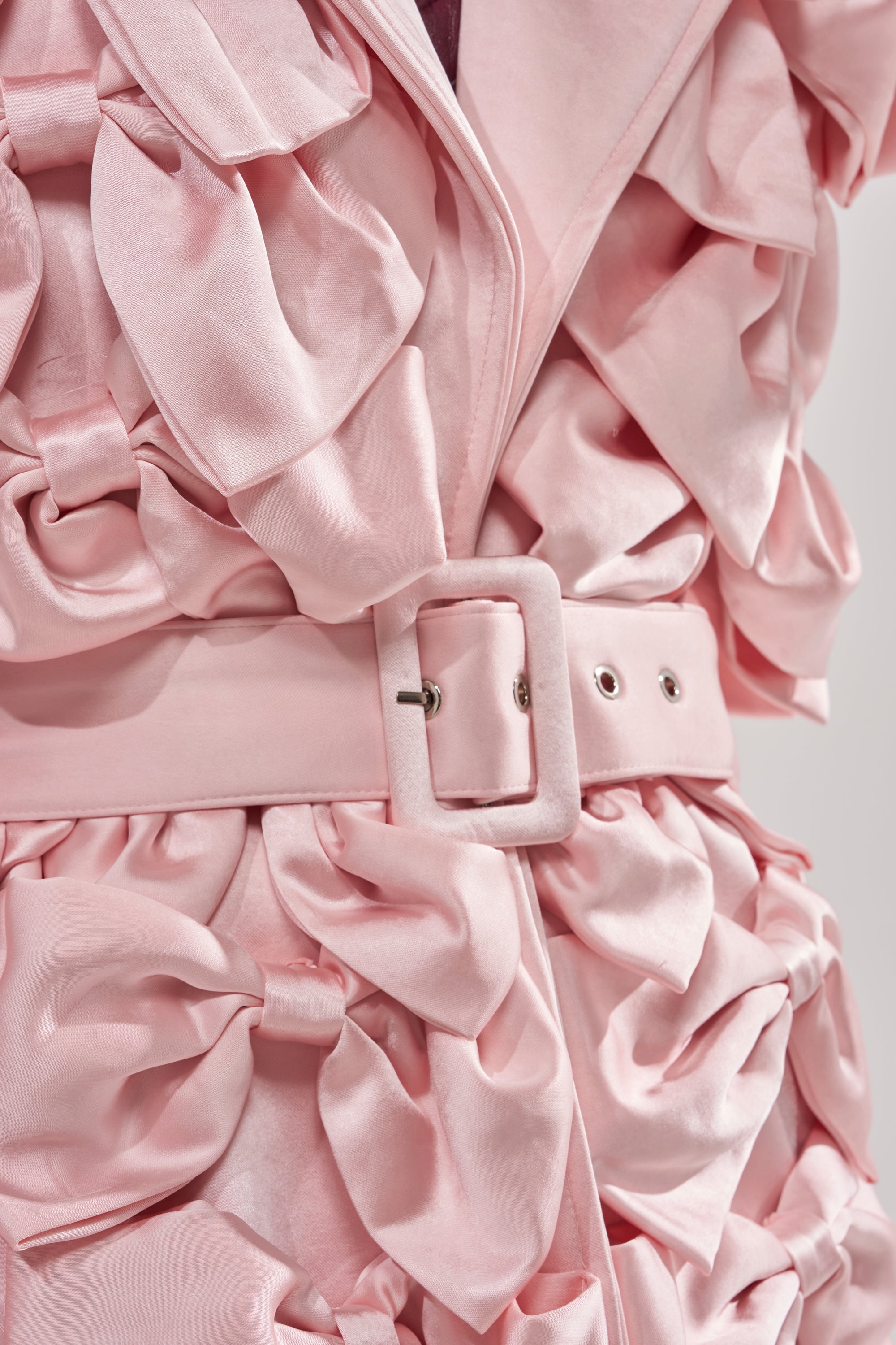 Close-up of the WRAP ME WITH A SATIN BOW TRENCH, a pink satin coat with large ruffled details and a matching belt with rectangular buckle. Its soft, shiny texture creates a luxurious, couture-inspired look.