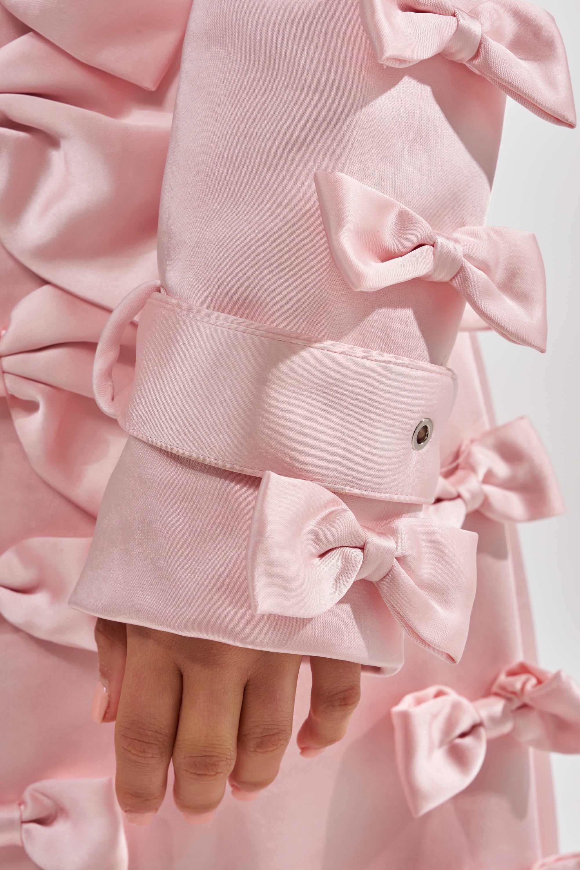A close-up of a hand at the side, wearing the WRAP ME WITH A SATIN BOW TRENCH in light pink, featuring smooth fabric and multiple pink satin bows, including on the sleeve cuff.