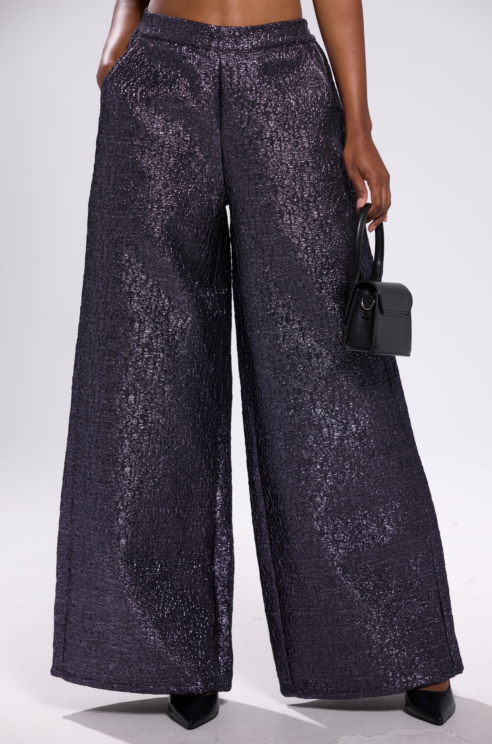 A person wears HIGH STANDARD BROCADE TROUSER—high-waisted, wide-leg, textured black sequins—with black pointed shoes and a small black handbag against a plain light gray background.