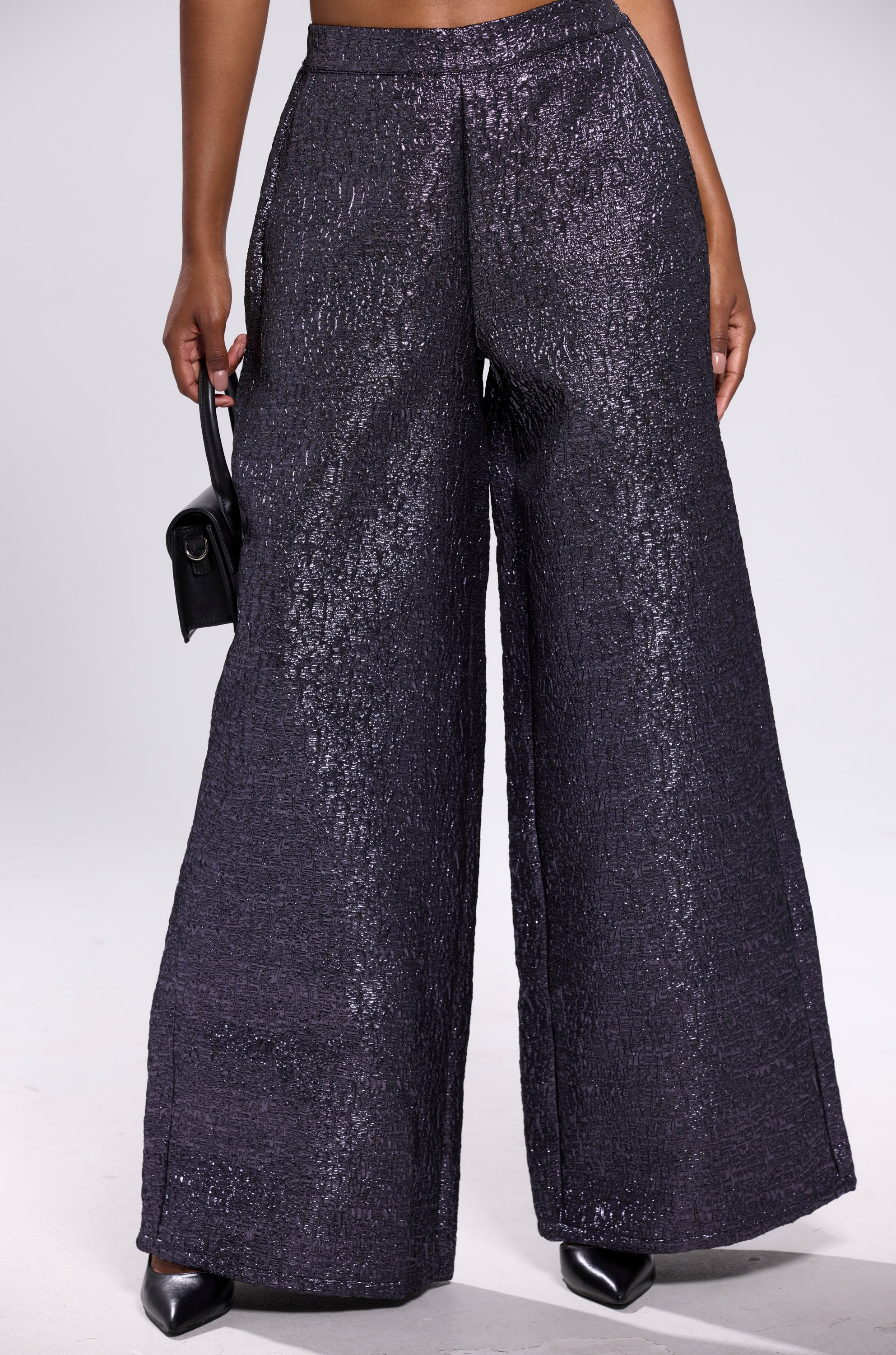 A person wears the HIGH STANDARD BROCADE TROUSER—wide-leg, high-waisted, and textured metallic—paired with a small black handbag and pointed black shoes, posed against a plain light background.