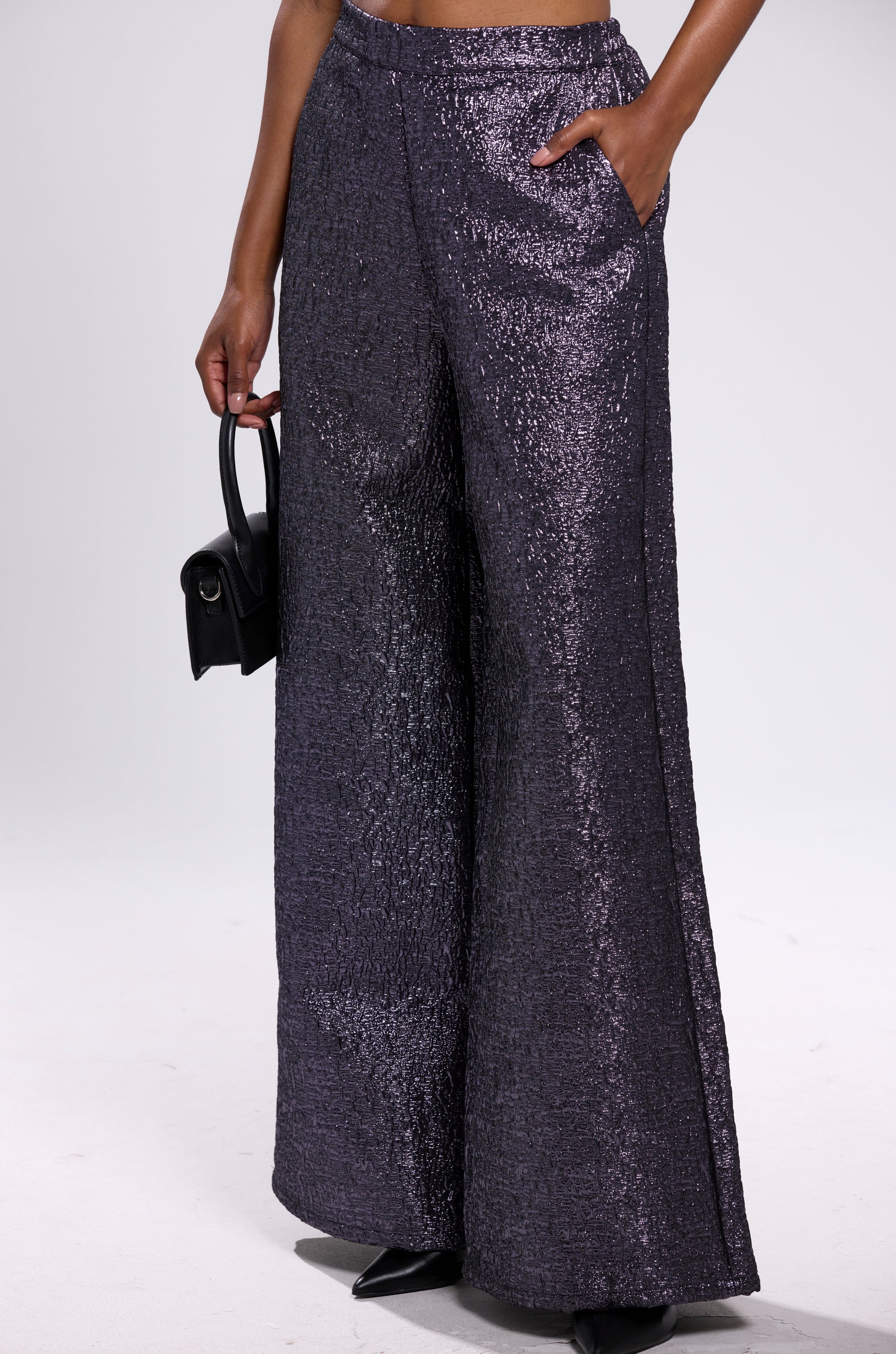 The lower body is shown wearing HIGH STANDARD BROCADE TROUSER in a dark purple metallic shade with a wide-leg fit, styled with a small black handbag, set against a plain light background.