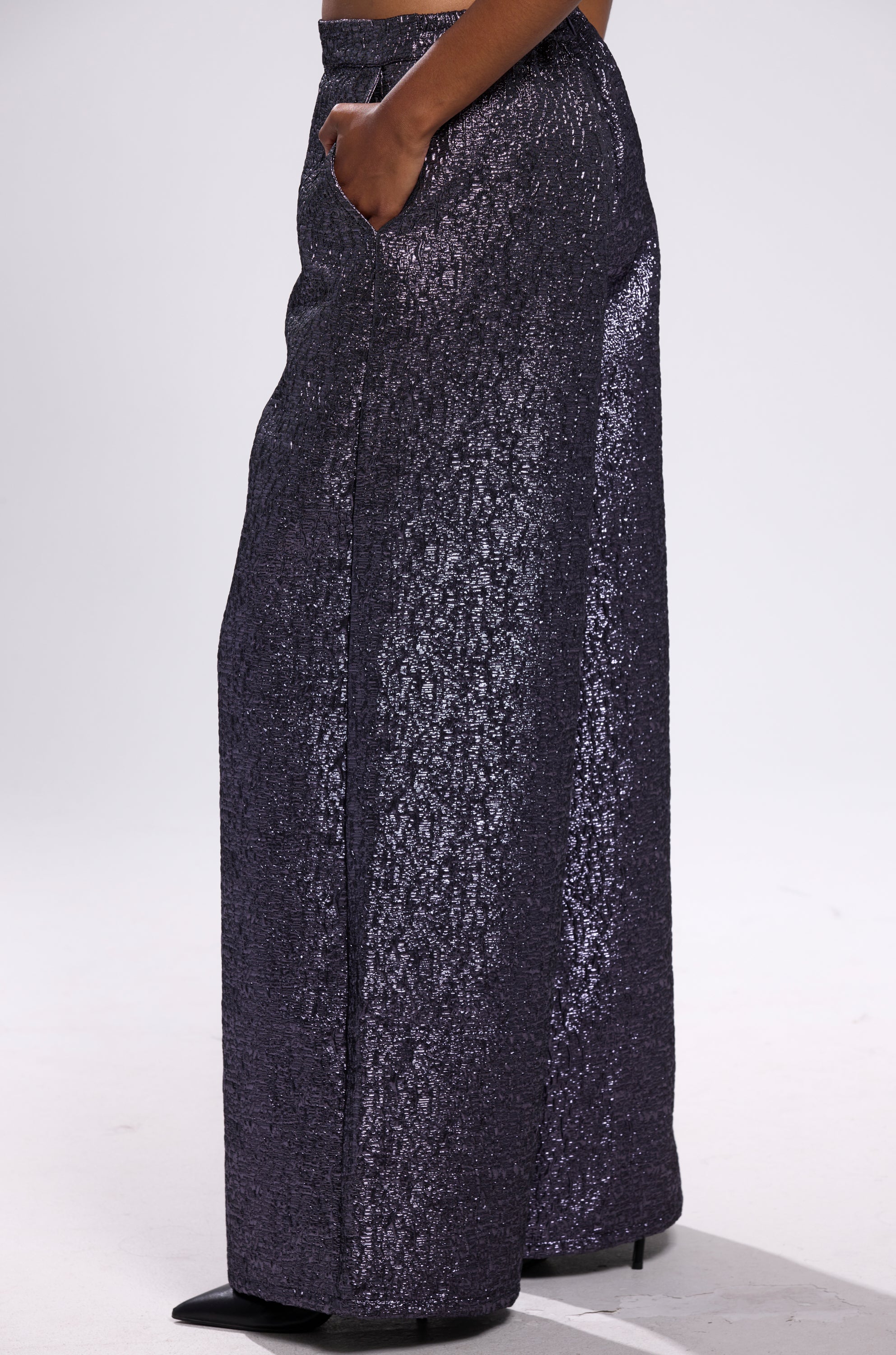 A person stands with hands in pockets, showcasing the HIGH STANDARD BROCADE TROUSER—high-waisted, wide-leg pants in sparkly black brocade—against a white background. Only their lower half is visible.