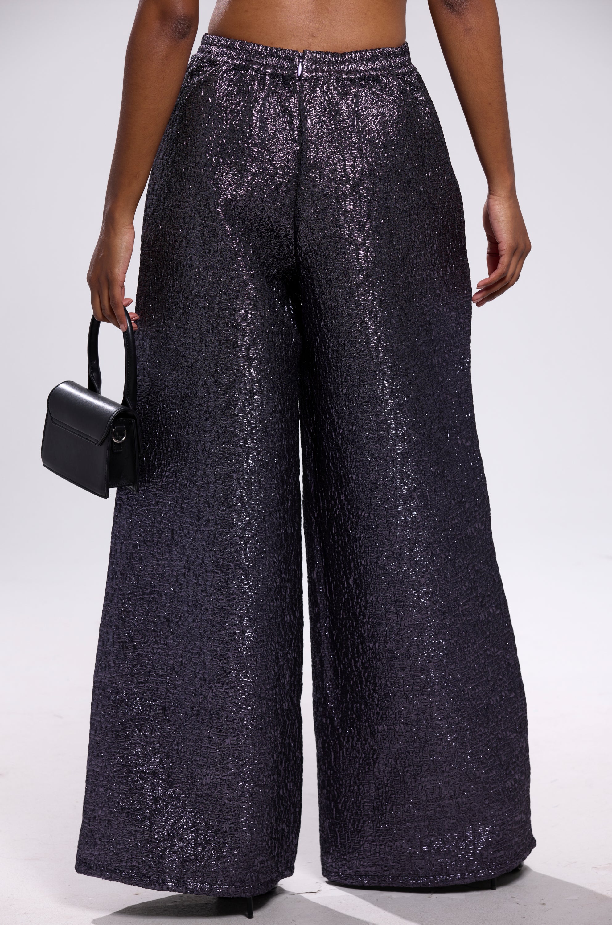 A person stands facing away, wearing HIGH STANDARD BROCADE TROUSER—high-waisted, wide-leg pants with textured black sequins—and holding a small black handbag against a plain, light-colored background.