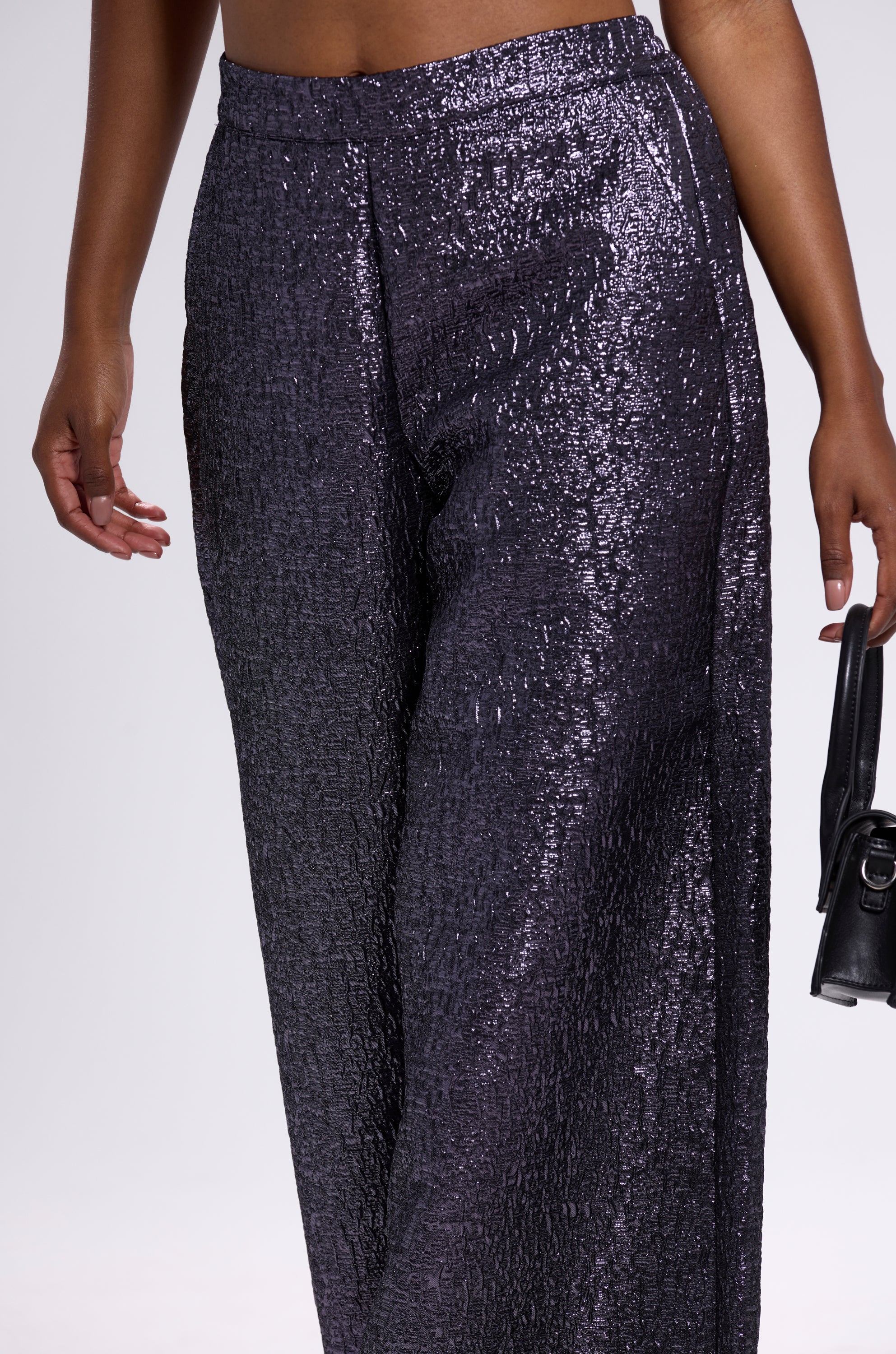 Wearing the HIGH STANDARD BROCADE TROUSER—high-waisted, wide-leg, metallic fabric—a person holds a small black handbag. The image is cropped to show the lower torso and legs against a plain background.