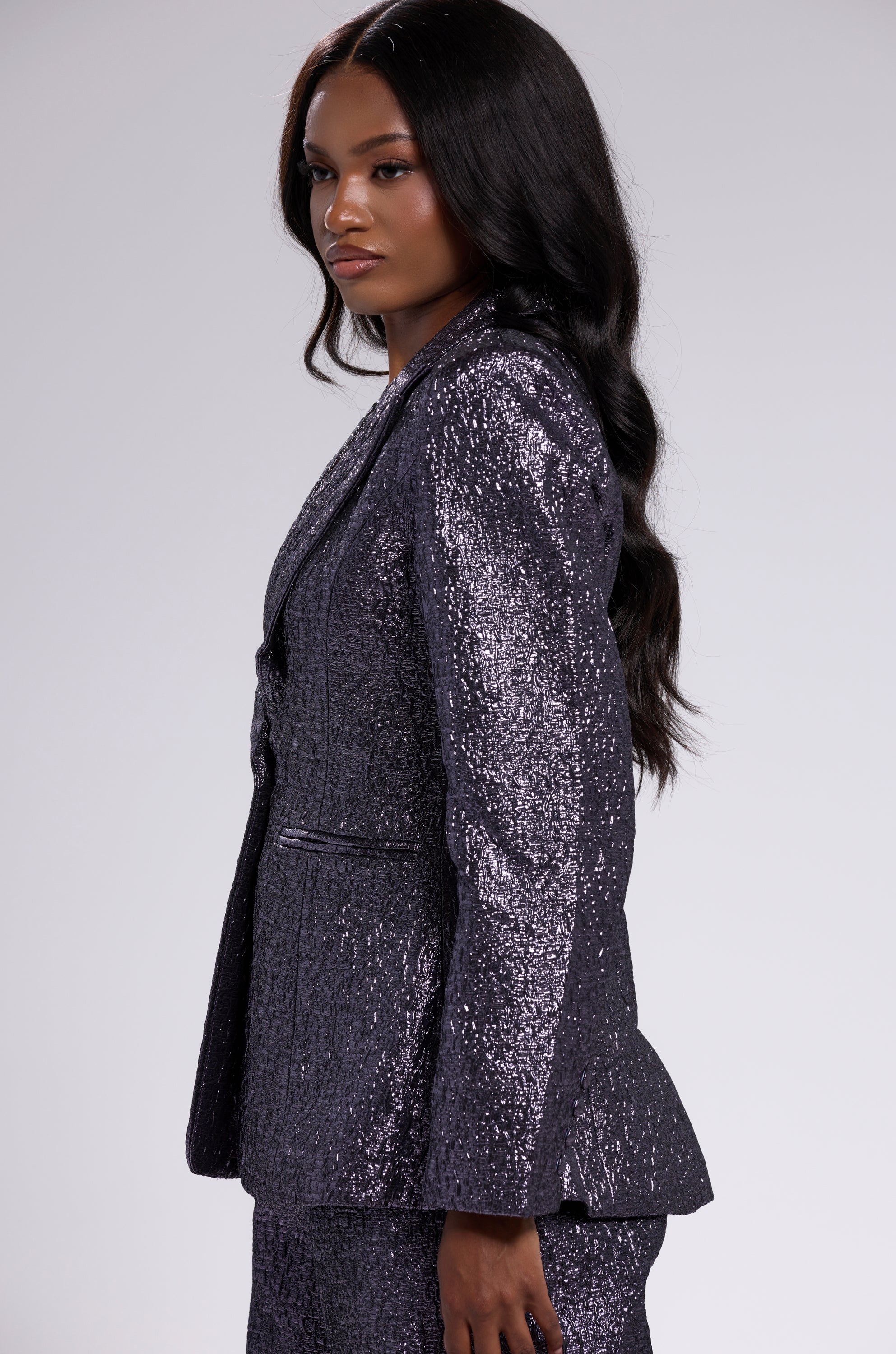 A woman with long, wavy black hair wears the HIGH STANDARDS BROCADE BLAZER and matching pants in shiny dark purple, standing in profile against a plain light gray background.