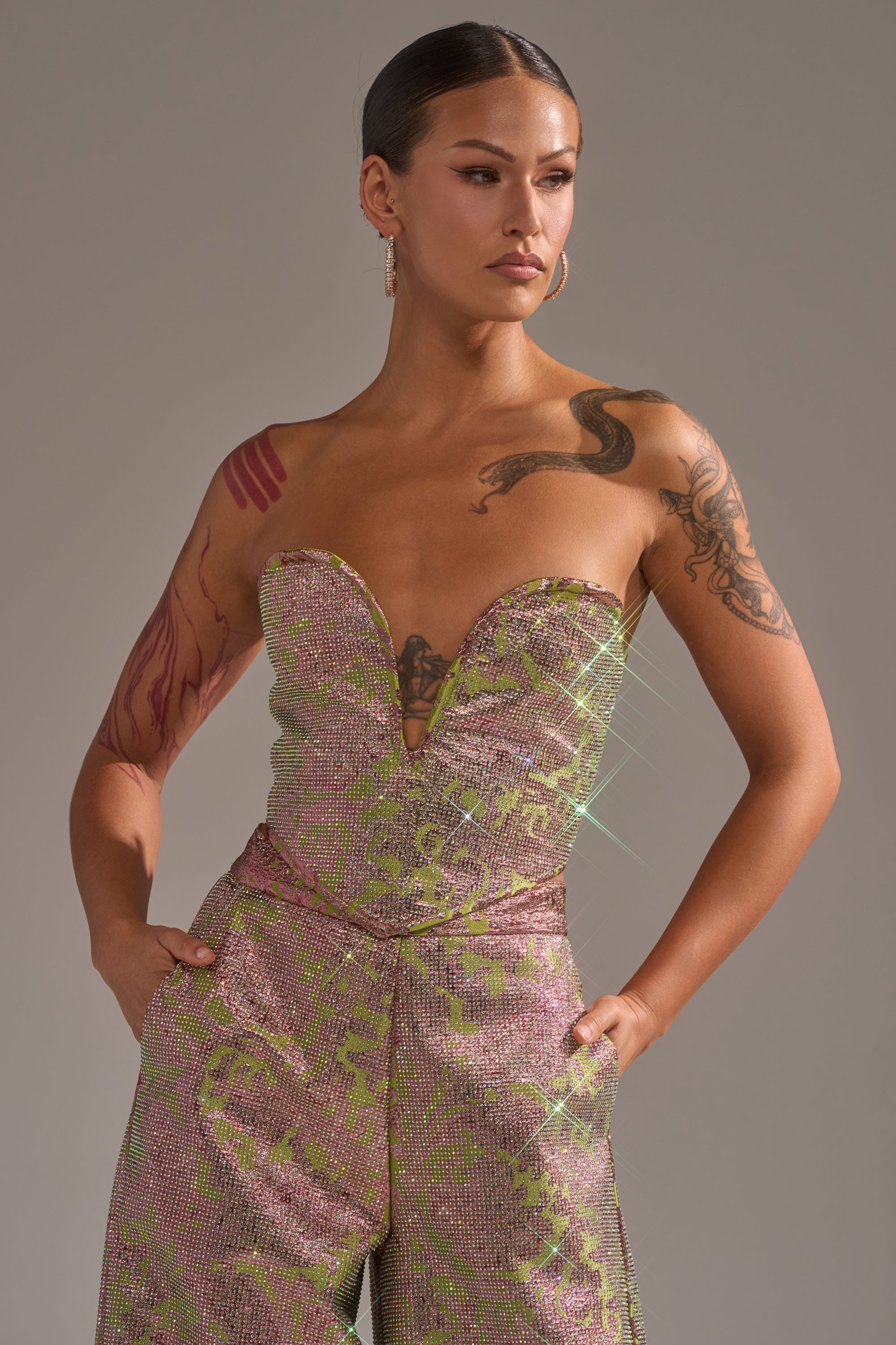 Wearing the TIME AND TIME AGAIN RHINESTONE CORSET, a person with tattoos, hoop earrings, and slicked-back hair poses confidently in a sparkling pink and green strapless jumpsuit with pockets and a deep neckline against a neutral background. 0