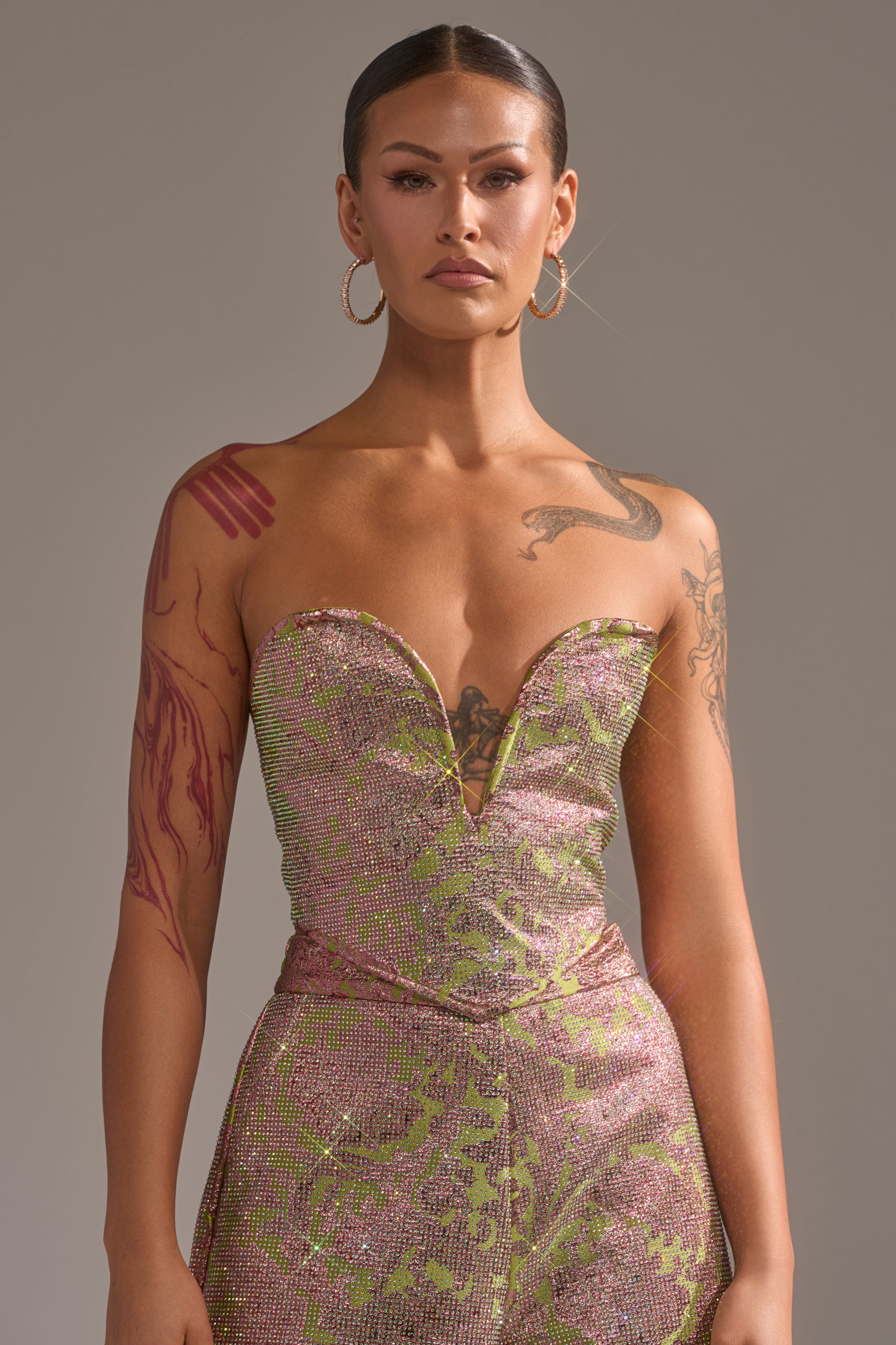 A person with slicked-back hair and hoop earrings wears the shimmering pink and green TIME AND TIME AGAIN RHINESTONE CORSET by AKIRA Label, showing arm and shoulder tattoos while posing confidently against a neutral gray background. 1