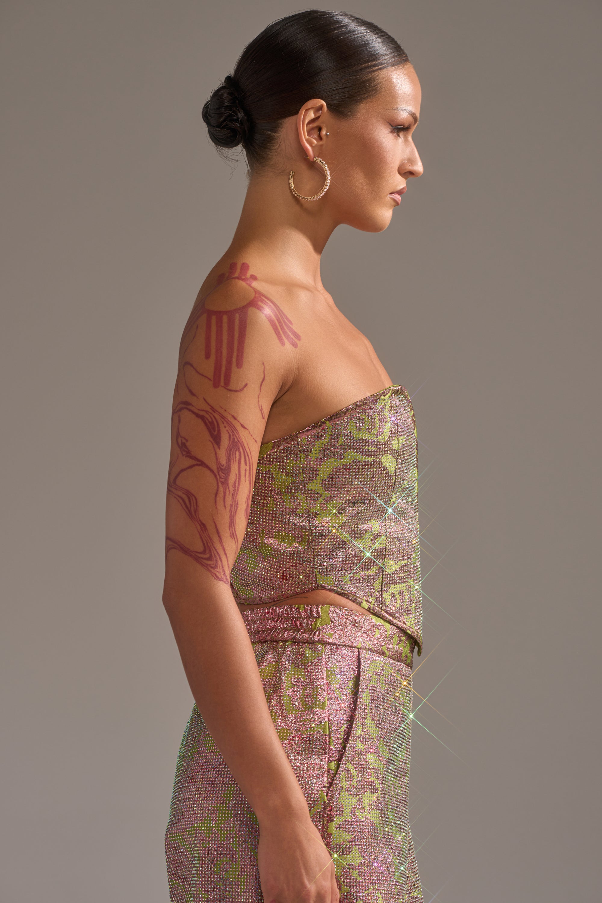 A woman with slicked-back hair in a low bun wears the TIME AND TIME AGAIN RHINESTONE CORSET and large hoop earrings. A bold red tattoo covers her upper right arm as she stands in profile against a neutral background, embodying AKIRA Label style. 2