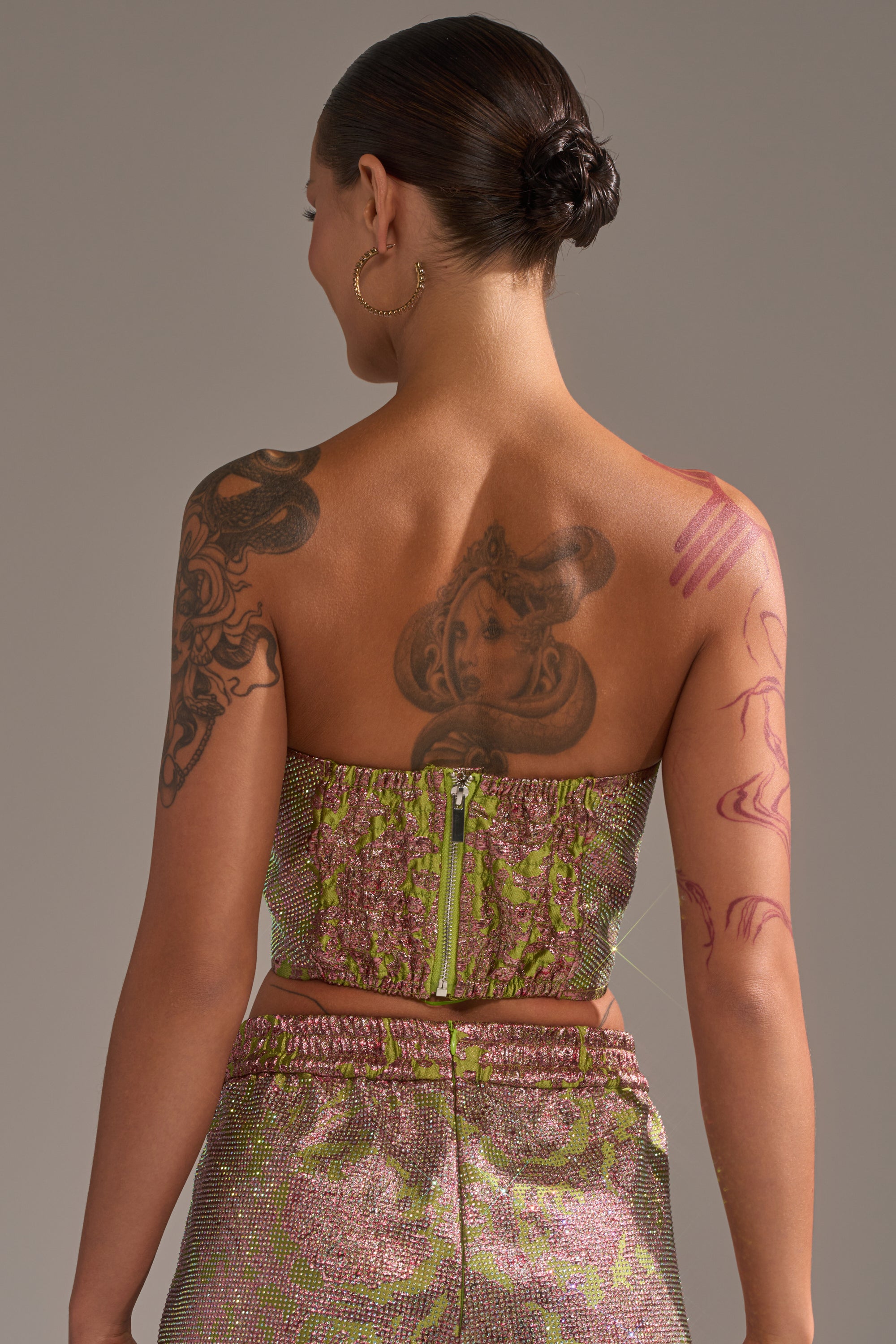 A person with tattoos on their back and arms is seen from behind, wearing the TIME AND TIME AGAIN RHINESTONE CORSET. Their hair is tied back in a neat bun, highlighting the corset’s sparkling rhinestone detail and elegant style. 3