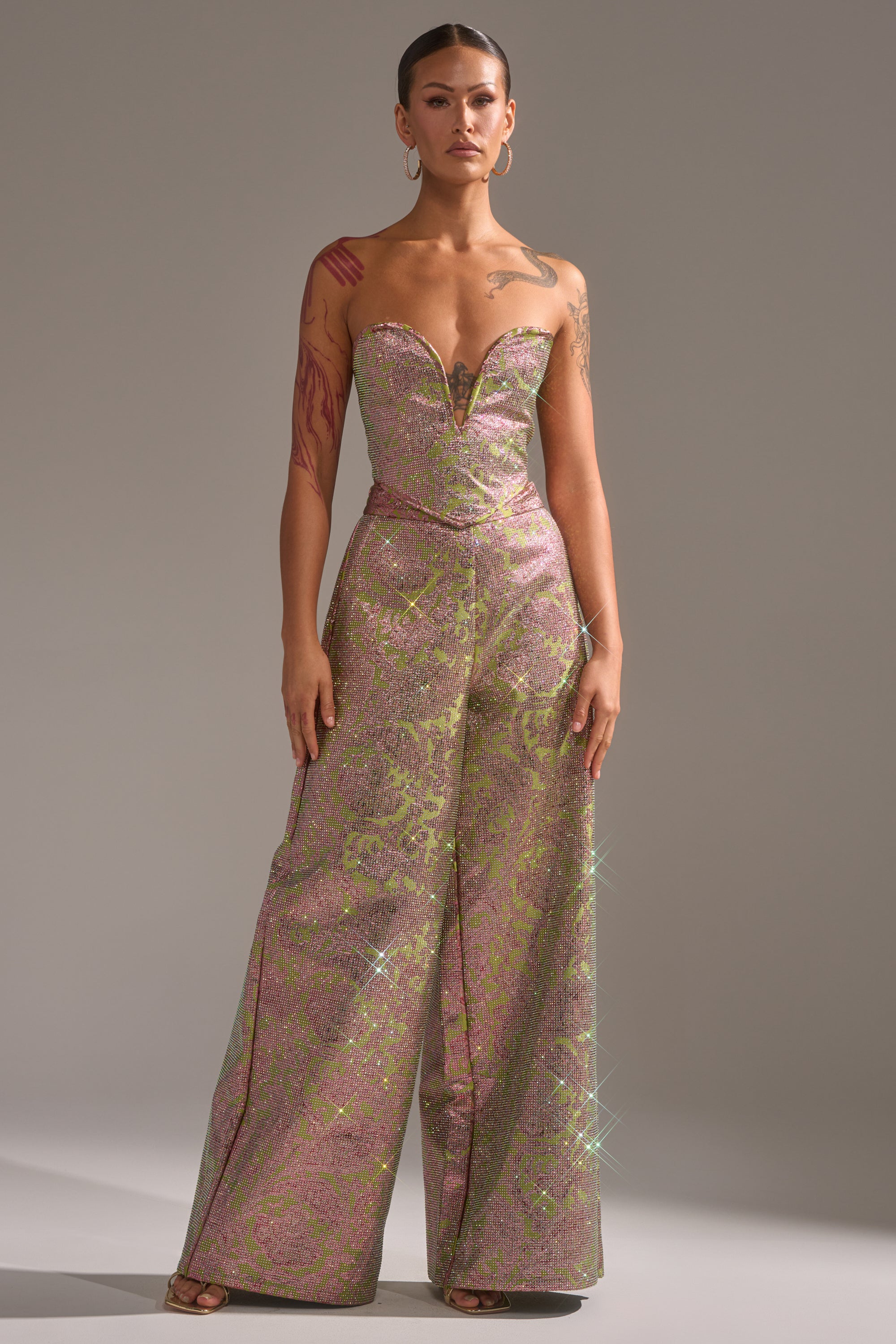 A woman wears the TIME AND TIME AGAIN RHINESTONE CORSET, a strapless, shimmery pink and green floral brocade piece with a plunging neckline, styled confidently against a plain gray background. 5